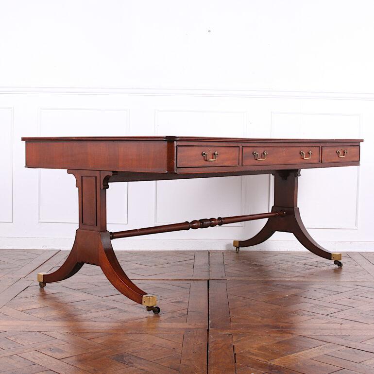 An English Georgian-revival double sided leather top library table or writing desk in mahogany, fitted with three drawers to each side and raised on a double-pedestal base with out-swept sabre legs with brass castors. The top with reeded edge and