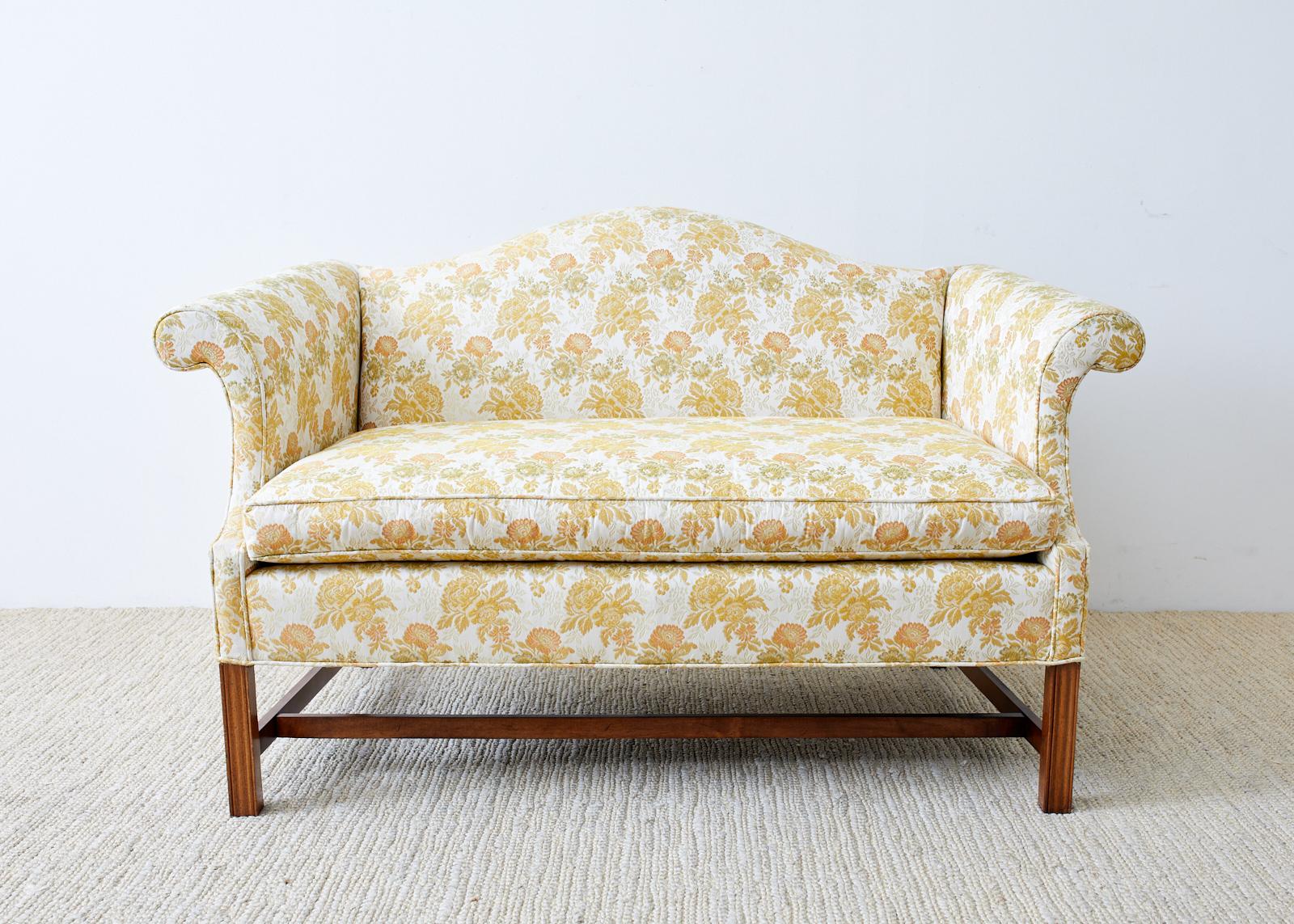 Charming English Georgian style settee or loveseat featuring a mahogany frame with a camelback hump form. The frame has a lovely thin arm that scrolls on each end giving the settee a classic profile. The seat has a thick, loose seat cushion that