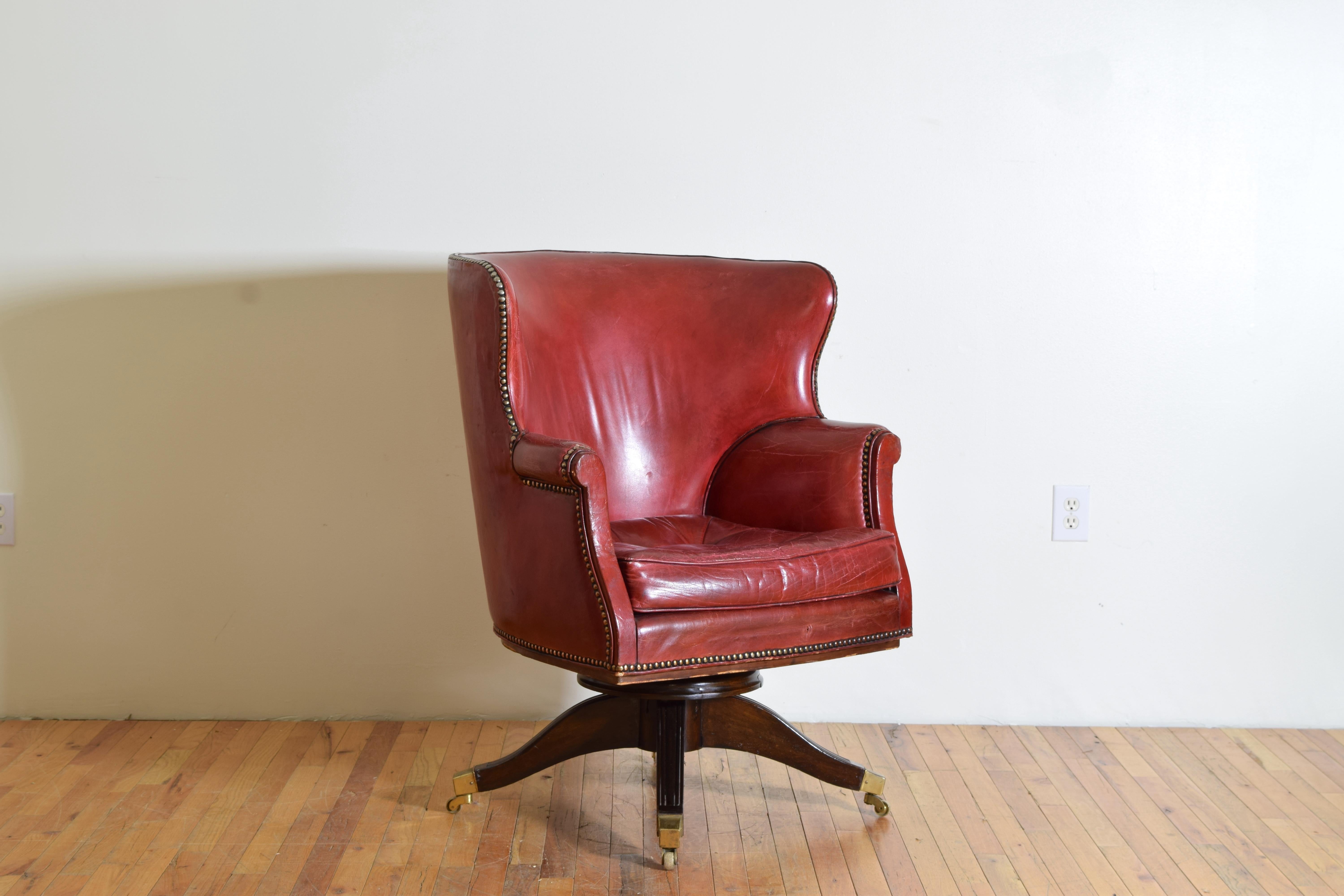 Having a concave backrest with wrap around wings, the leather trimmed in brass nailheads, with tufting to the backrest, raised on fluted and splayed mahogany legs ending in brass casters