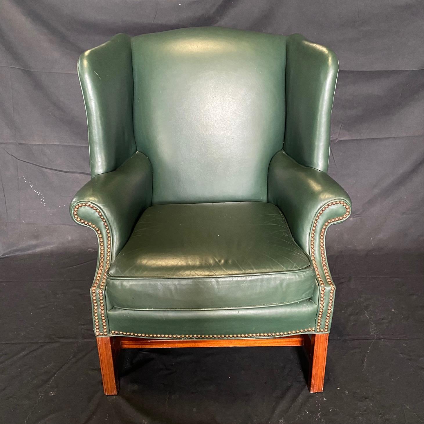 Beautiful and comfortable mid century leather wing chair featuring rich green leather upholstery. Made in the grand English Georgian style with a mahogany hardwood frame. The chairs have large wings conjoined to rolled arms with decorative brass