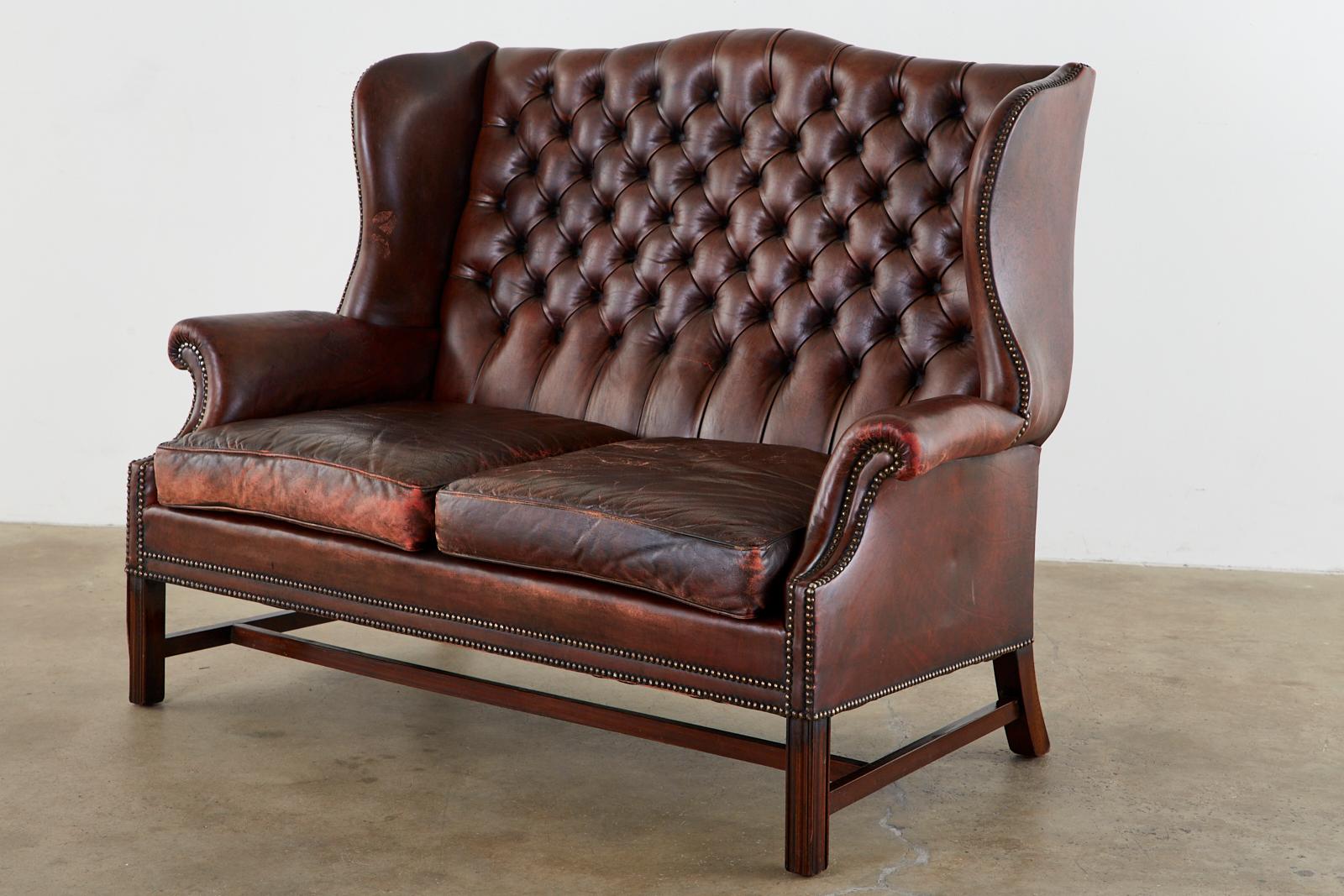 Beautifully aged English Chesterfield wingback settee or sofa. Featuring a tufted cordovan leather upholstery bordered by brass tack nailheads. The mahogany frame made in the Georgian taste features English rolled arms and large fully developed