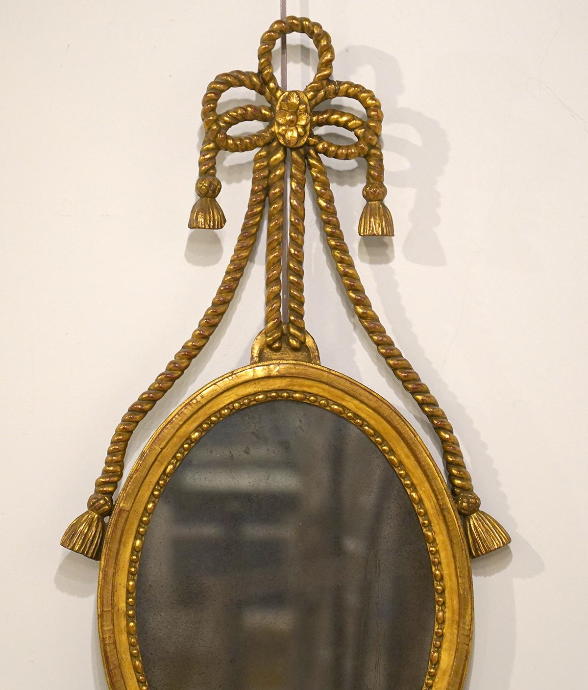 This rather rare Georgian wall mirror features an oval mirror in a beaded molded frame supporting an elegant large bow on the top from which ropes and tassels are gracing the miror frame. The bottom is adorned by ribbons and drapes centering a