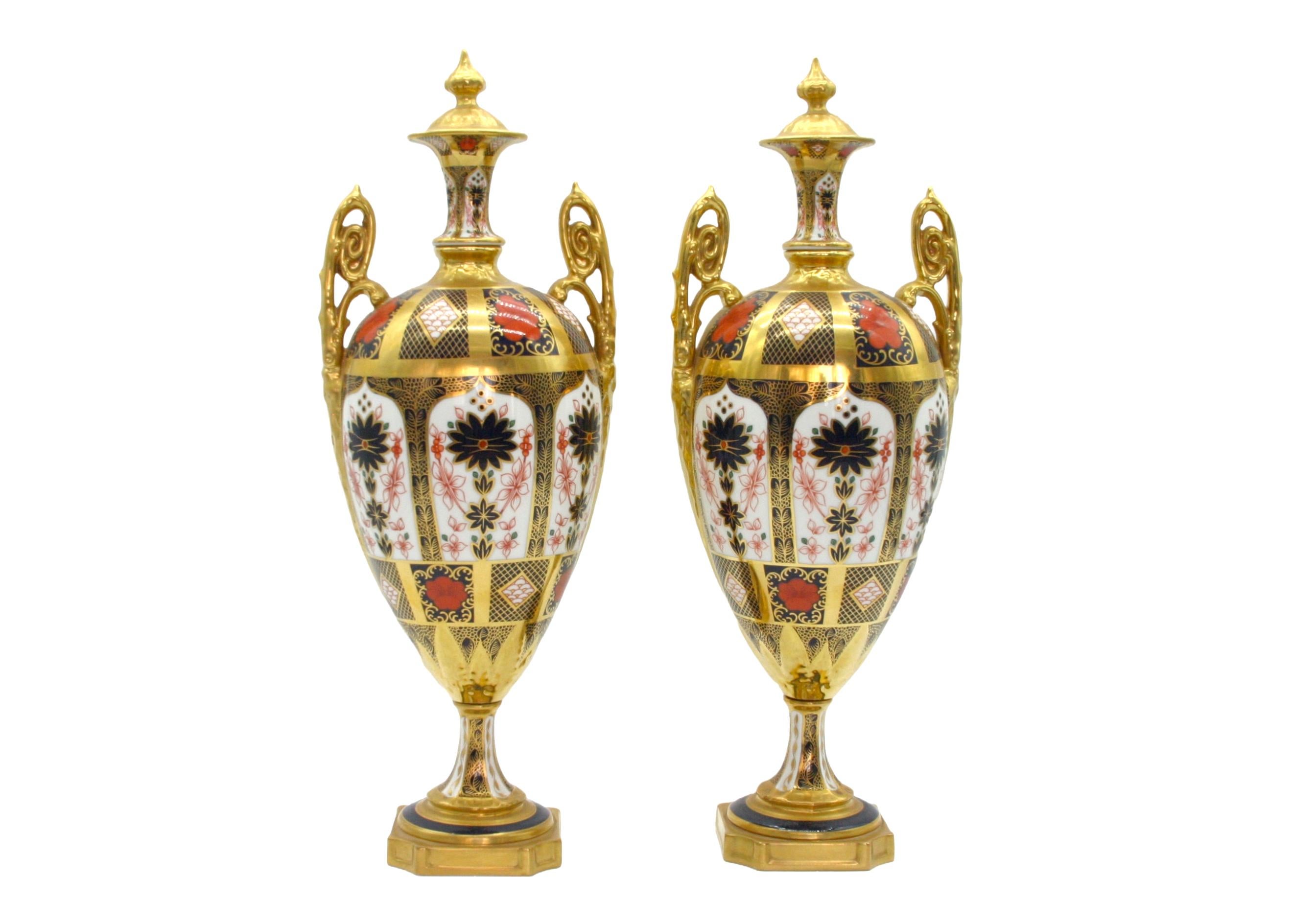 Beautifully gilt and hand painted Royal Crown Derby pair Imari pattern porcelain decorative urns/vases with gilt twin side handles. Each urn features an exterior 22k gold and handpainted details with an attached gilt gold cover and raised on