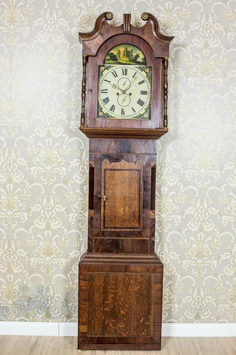 We present you this grandfather clock with an oak case, which is partially veneered with mahogany, with inserts made of fillet stripes.
The small roof of the case has small columns that flank the door, and is topped with an irregular, volute