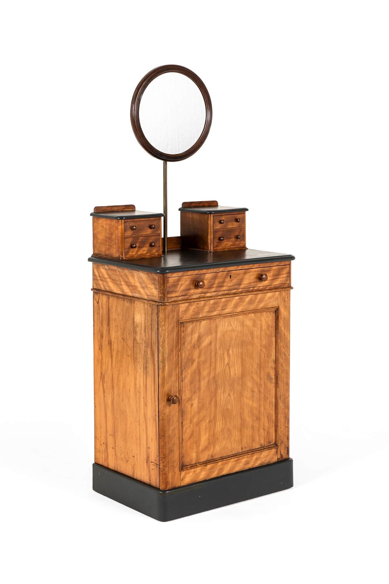 An unusual late Victorian gentlemen’s dressing stand in satinwood by Heal and Son.

The top has an arrangement of four small drawers in two separate banks flanking a circular mirror on a brass pole upright.

The mirror is articulated by a bolt to