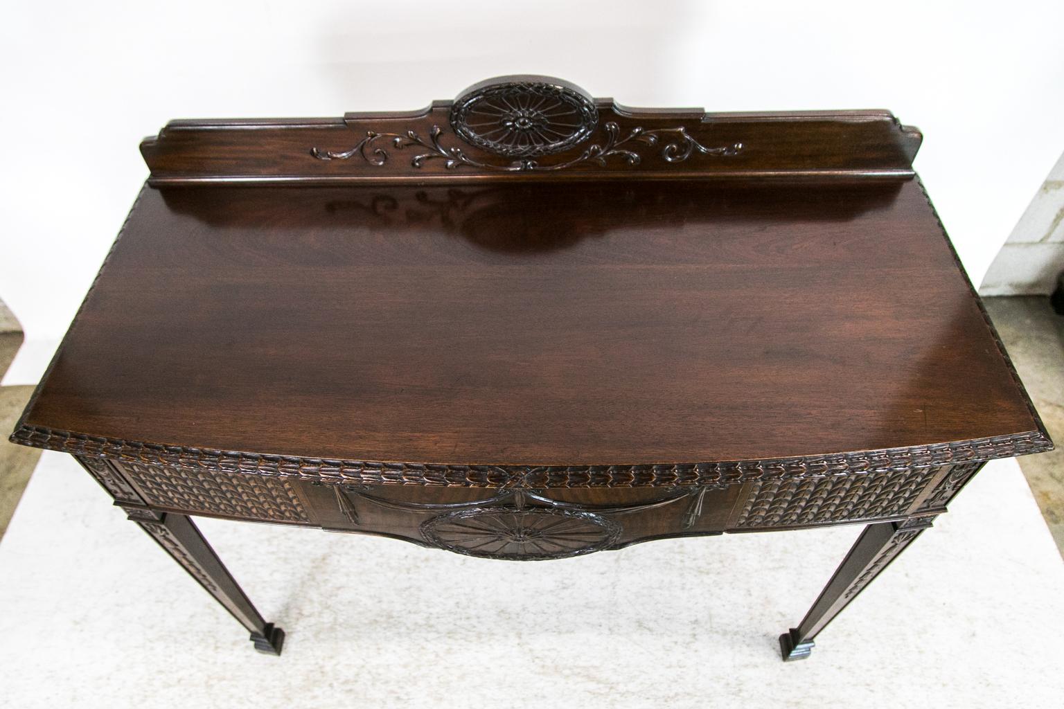 The rear gallery of this console table has a carved oval crest above carved leaf arabesque. The top molding is carved with a repeating stylized bell flower. The apron consists of a center door carved with ribbon swags supporting a carved oval