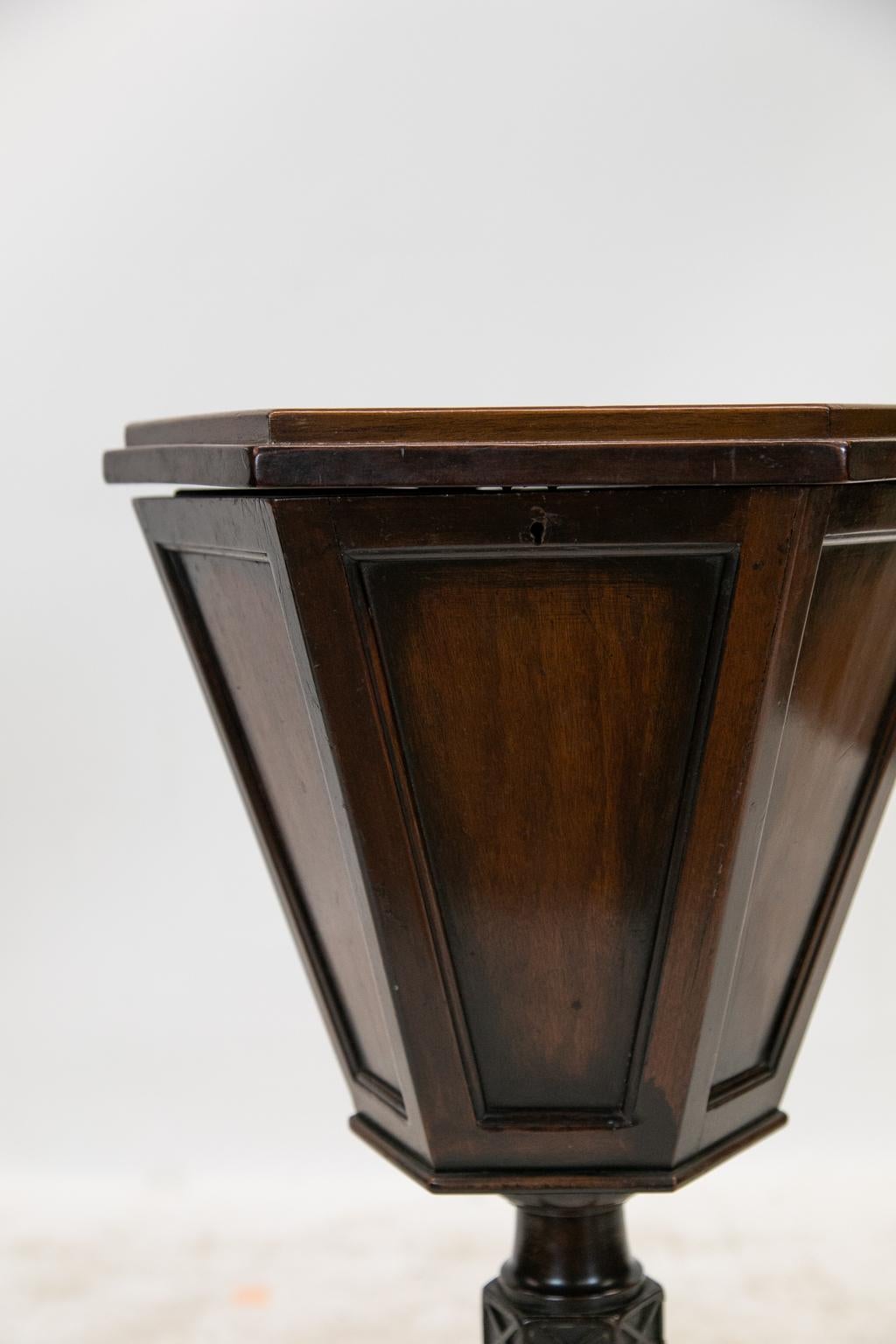 The top of this English mahogany hexagonal sewing table has a bullnose gallery. The body is in a trapezoid shape. The six sides have recessed panels with molded edges. The stem has a carved blind fretwork hexagonal knob. The tripod legs have carved