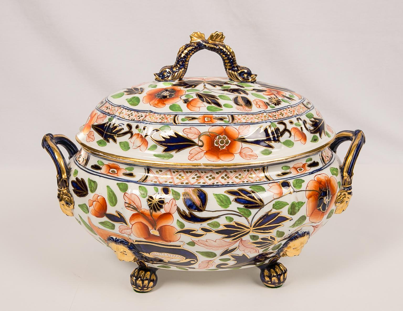 A large antique English porcelain soup tureen and stand decorated with a colorful Imari design showing an overflowing vase on the garden terrace. This tureen and stand has eye-catching deep cobalt blue finials, handles, and trim which are decorated