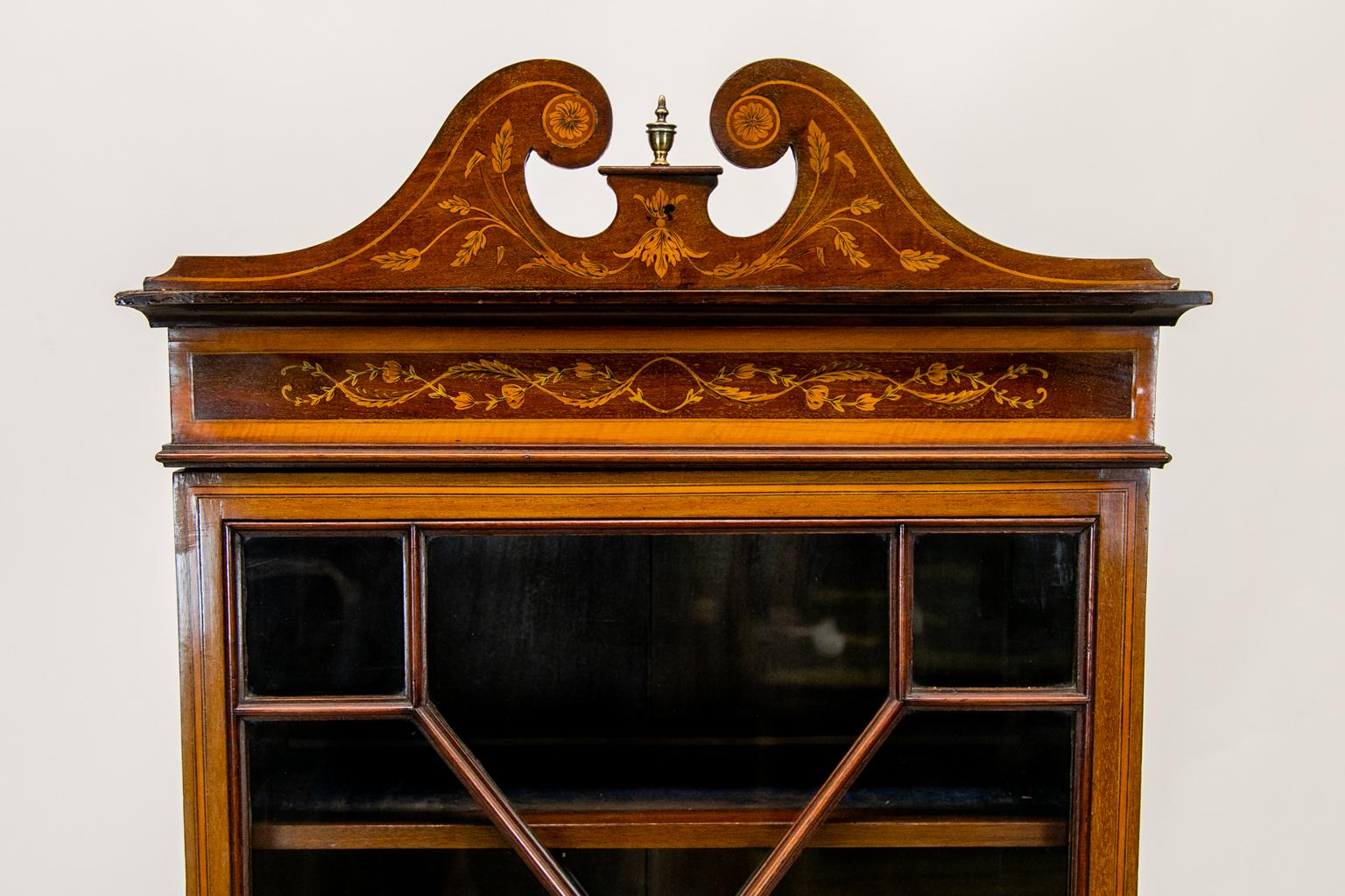 This English inlaid bookcase has an eight pane astragal door which is banded with satinwood and ebony inlay. It has the original working lock and key. The cornice has a broken arch pediment inlaid with floral arabesques with a small beaded brass