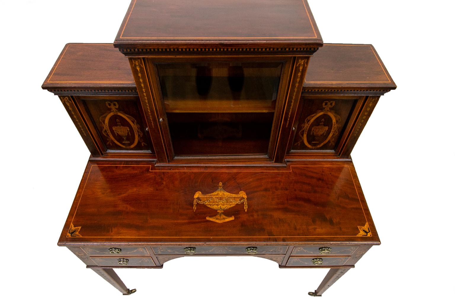 This ladies desk is intricately inlaid with classical urns, draped ribbons and bellflowers on the stiles and legs. There is boxwood line inlay on the top, drawers, legs, and sides. The center cabinet has a beveled glass door with molded framing. The