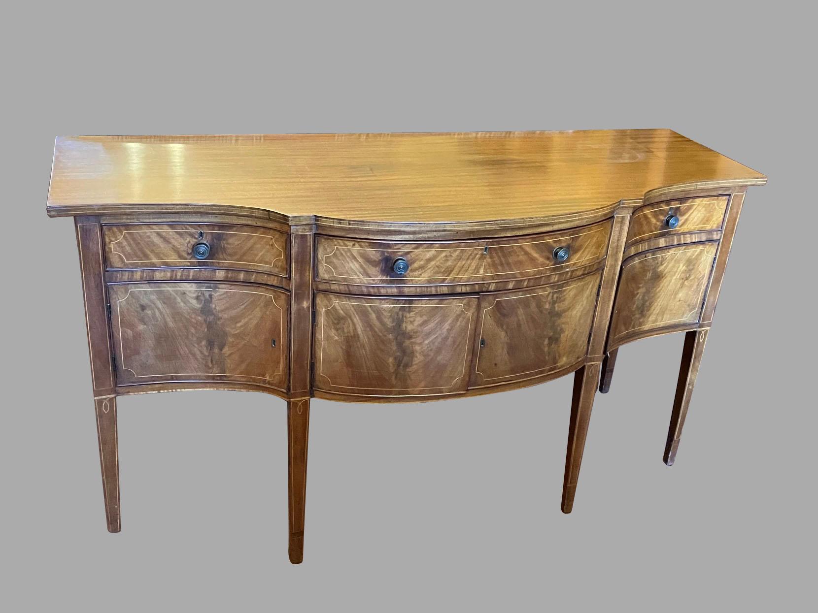 An English Hepplewhite style sideboard of serpentine form with nicely figured matched veneers, one long and two short drawers over 3 cabinet doors, supported on square tapered boxwood line inlaid legs. Circa 1900.