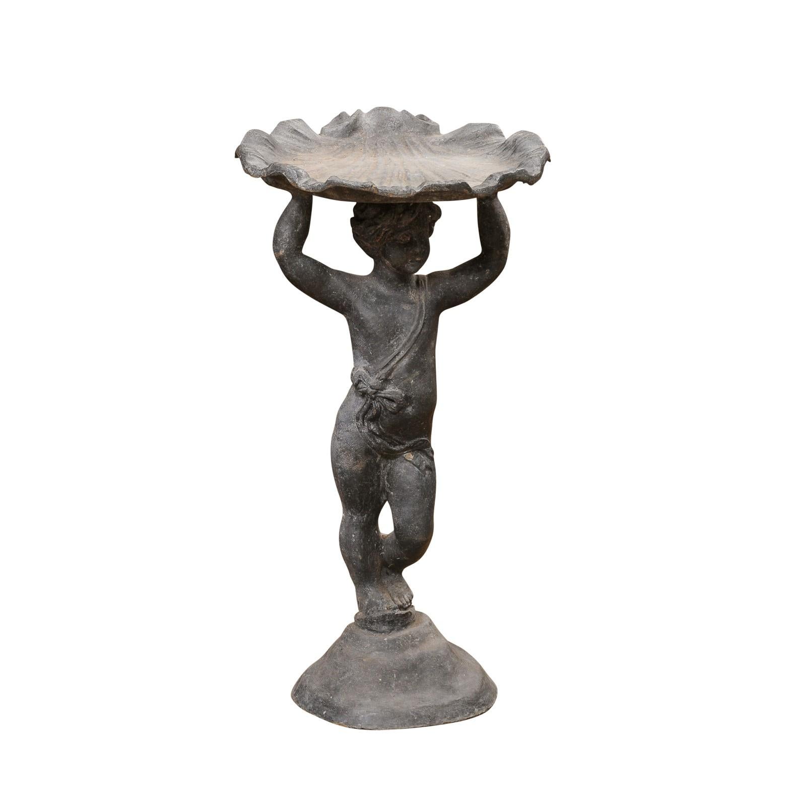 An English Greco-Roman style iron bird bath from the 20th century depicting a standing cherub carrying a sea shell on his head. Experience the enchanting fusion of English craftsmanship and Greco-Roman artistry with this 20th-century iron bird bath,