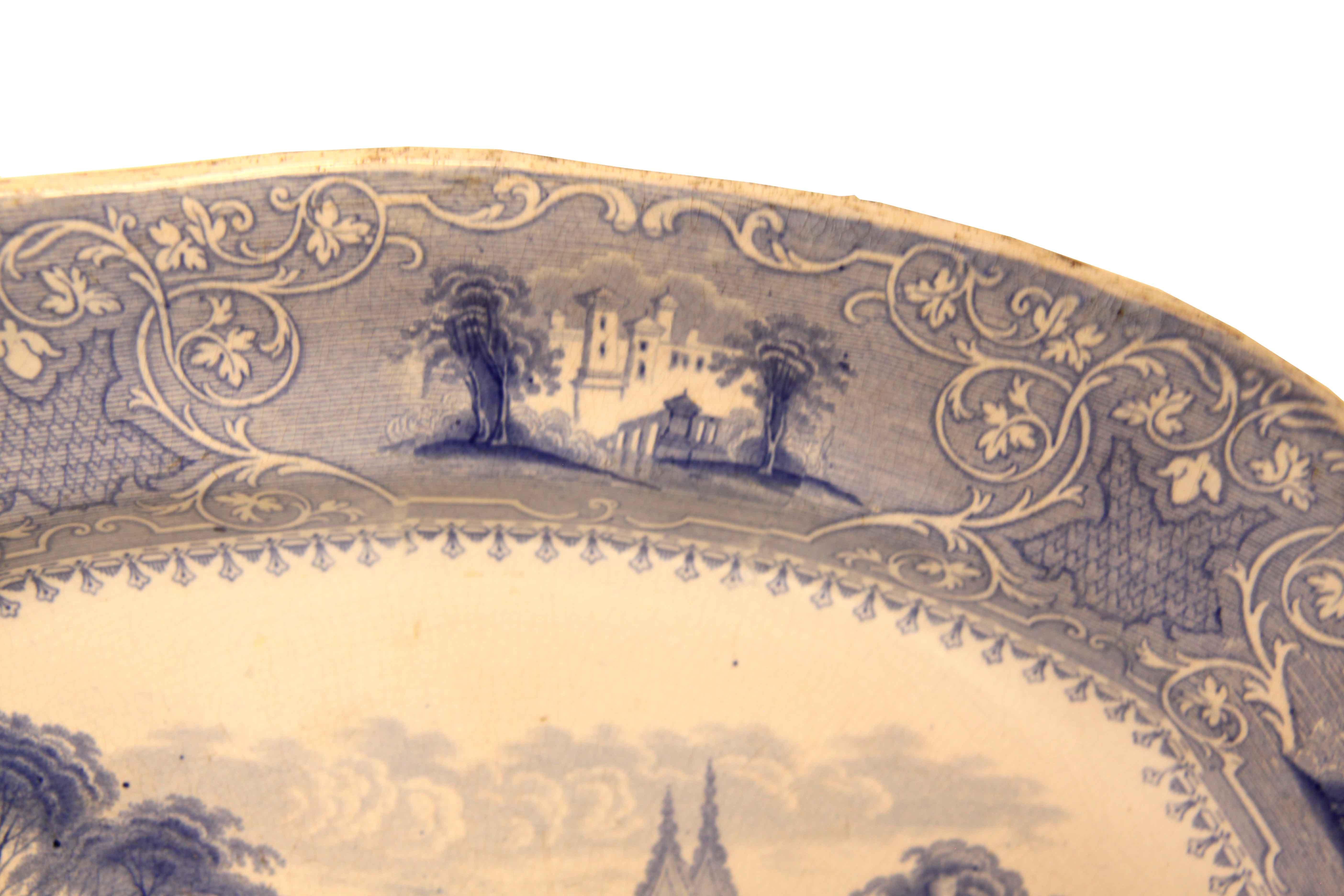 English ironstone blue and white platter from the Staffordshire region, the border features alternating scenes of castles with tress and stylized foliate and arabesques.  The main body depicts a river scene with ladies conversing and a fountain