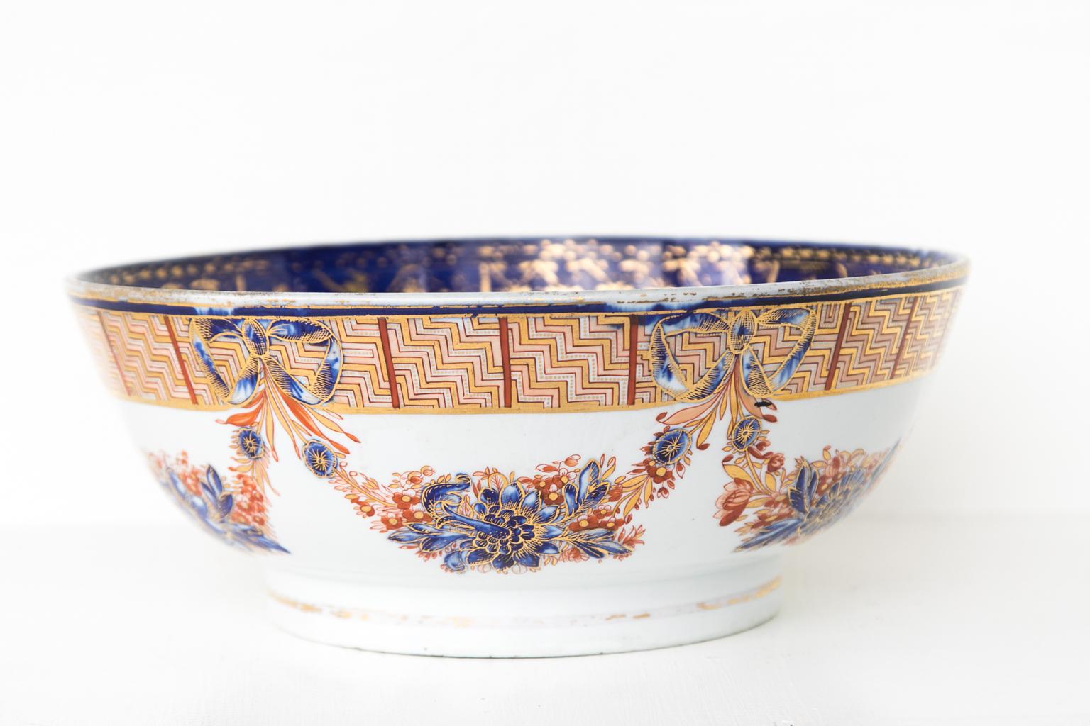 English ironstone bowl, featuring a cobalt blue interior rim with gilt highlights and a center medallion surrounded by leaves in the center of the interior. The exterior has red and blue floral swags connected with bows.
    