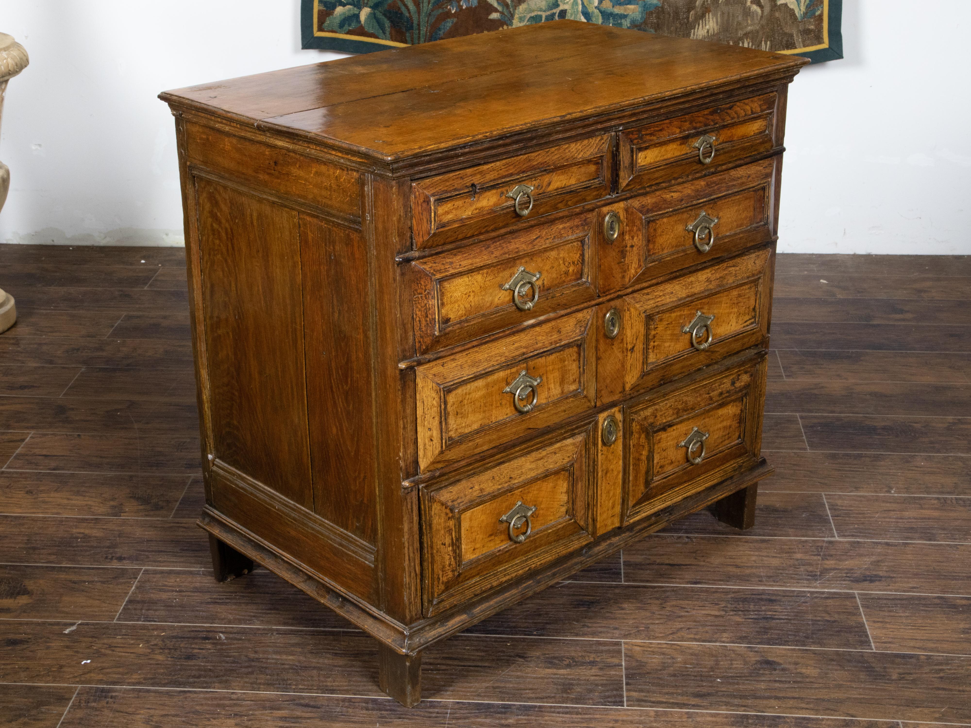 An English George III period oak commode from the early 19th century, with five graduating drawers, raised panels, brass hardware and short feet. Created in England during the George III period in the early years of the 19th century, this oak