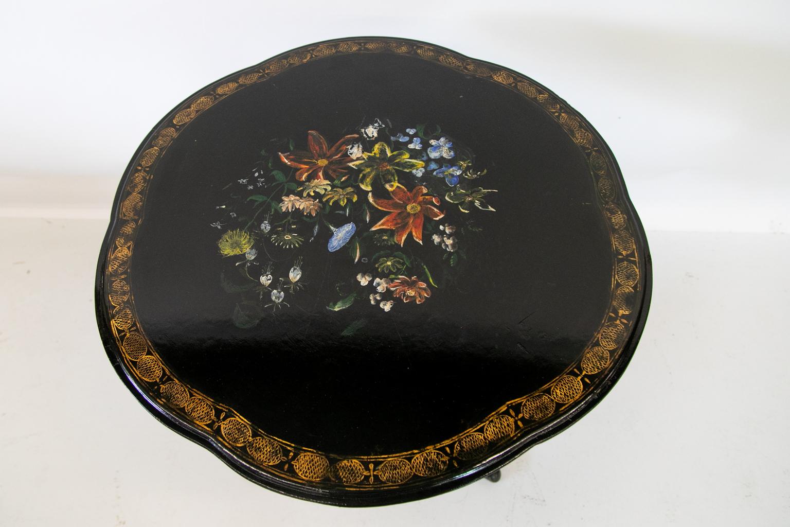 English lacquer tilt-top table has a scalloped top with a gold simulated pinecone border surrounding a large floral hand painted center. There are gold embellishments on the base.
    