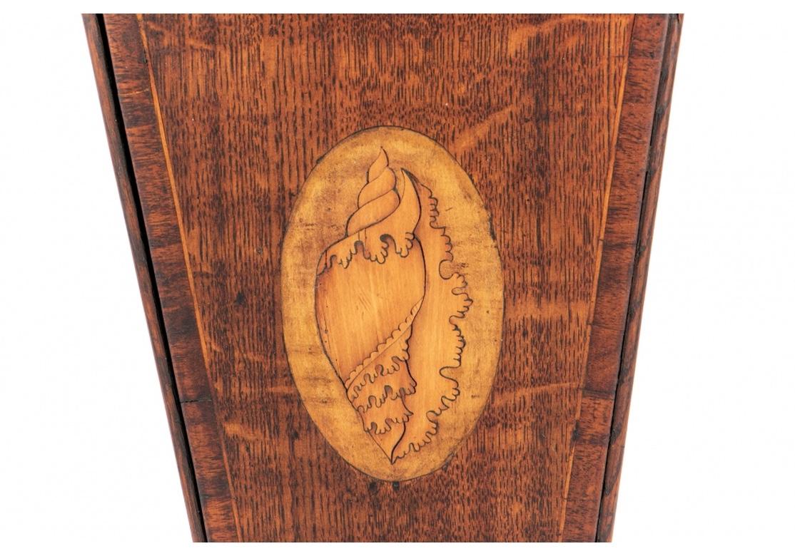 A tall tapering form in oak with a shaped carved back and hinged top. The front panel with a fine satinwood inlaid shell.
Measures: Height 18 3/4
