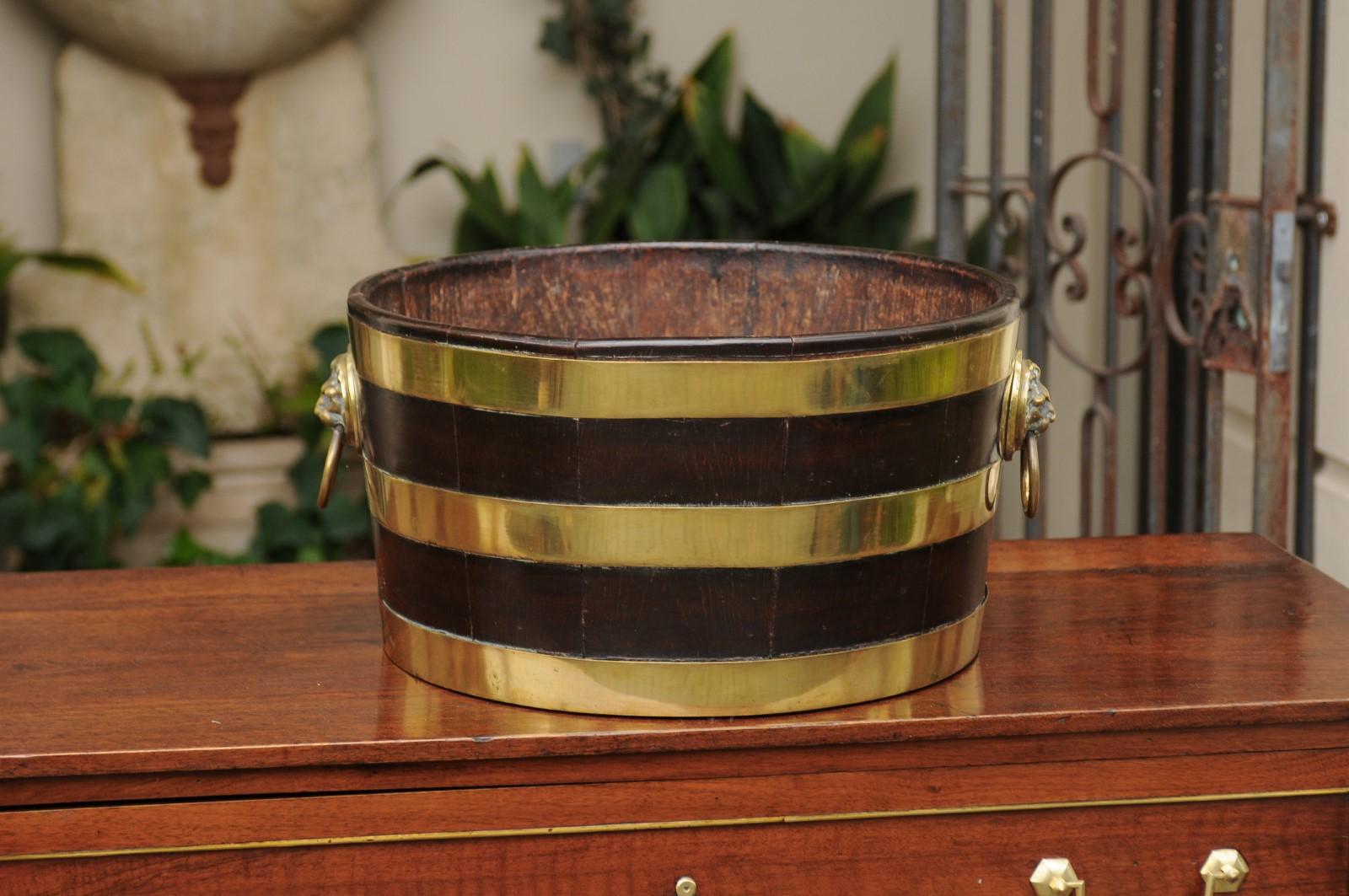 An English round oak wine cooler from the late 19th century with brass accents and lion handles. This lovely English wine cooler features a circular oak structure, strengthened on the outside by three brass horizontal braces. The cooler showcases