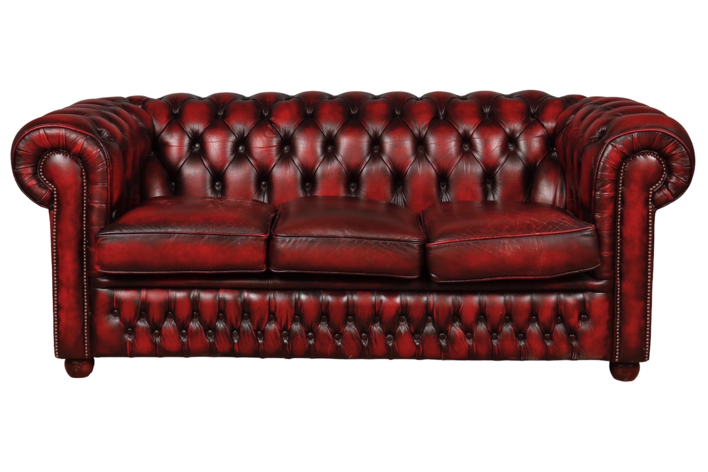 An English three seater Chesterfield sofa upholstered in oxblood red leather.  Diamond tufted on the back and front skirt, secured with a brass nailhead trim. Finished with wooden bun feet.
