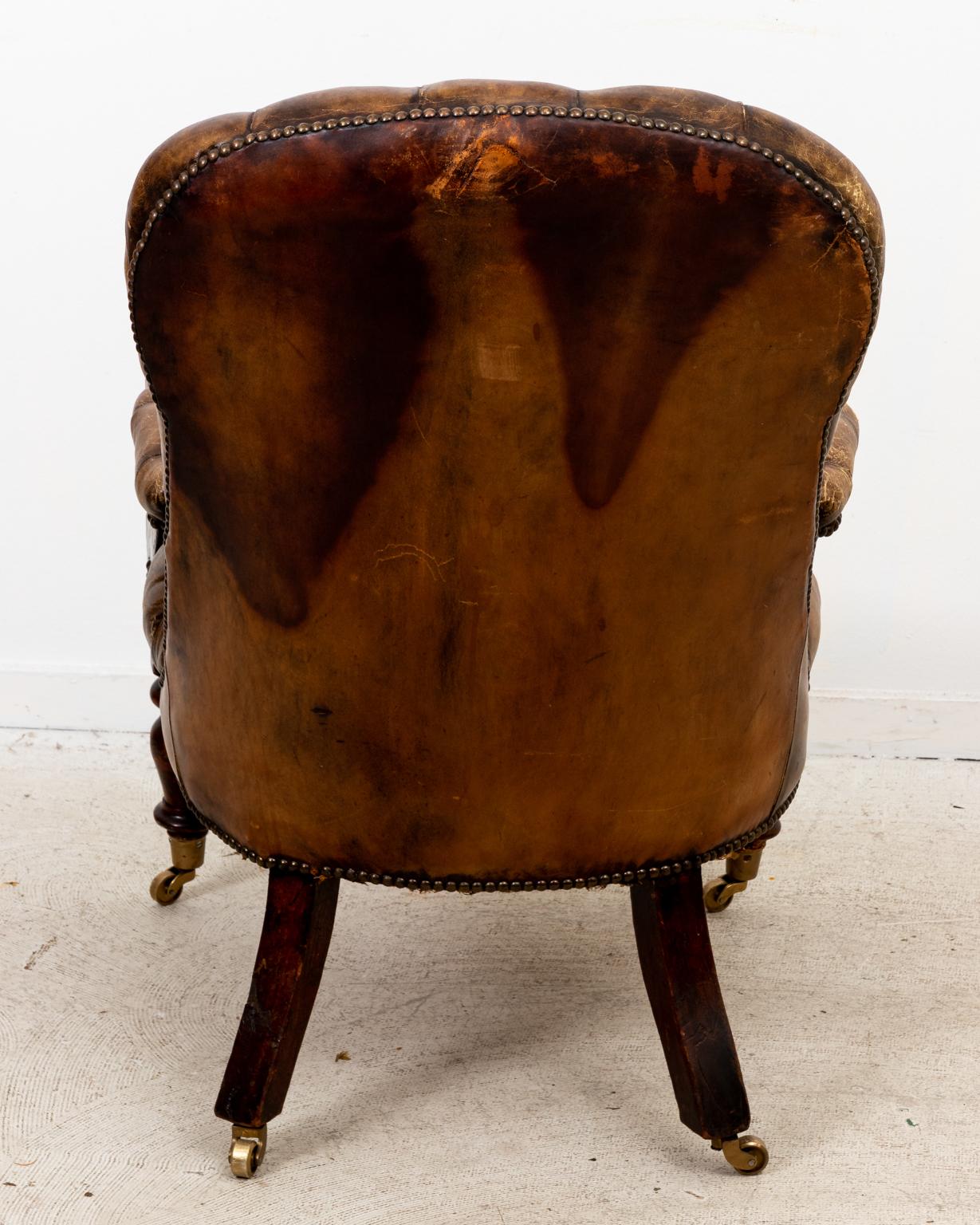 Circa 1830s English tufted leather library chair with carved, scrolled arm holds in the William IV style on castors. Made in England. Please note wear consistent with age.