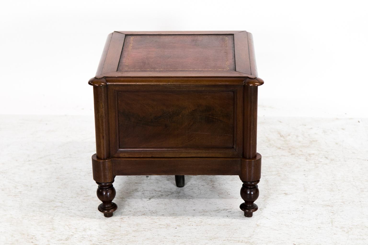 This mahogany leather top commode has turned front columns that terminate in turned legs. The front has a recessed flame mahogany panel framed with shaped molding. The top has an inset panel with gold and blind tooled leather. The interior retains