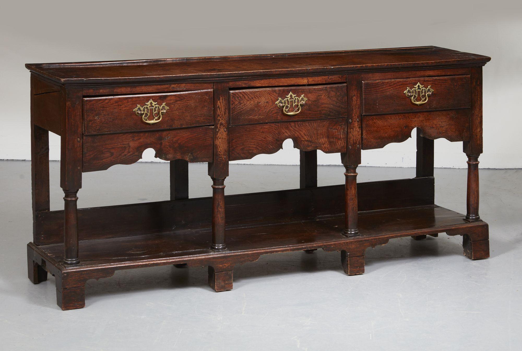 An 18th century English oak low dresser having three drawers with scalloped apron and column turned legs over potboard shelf. Desirable small proportions, elegant form and wonderful patina.