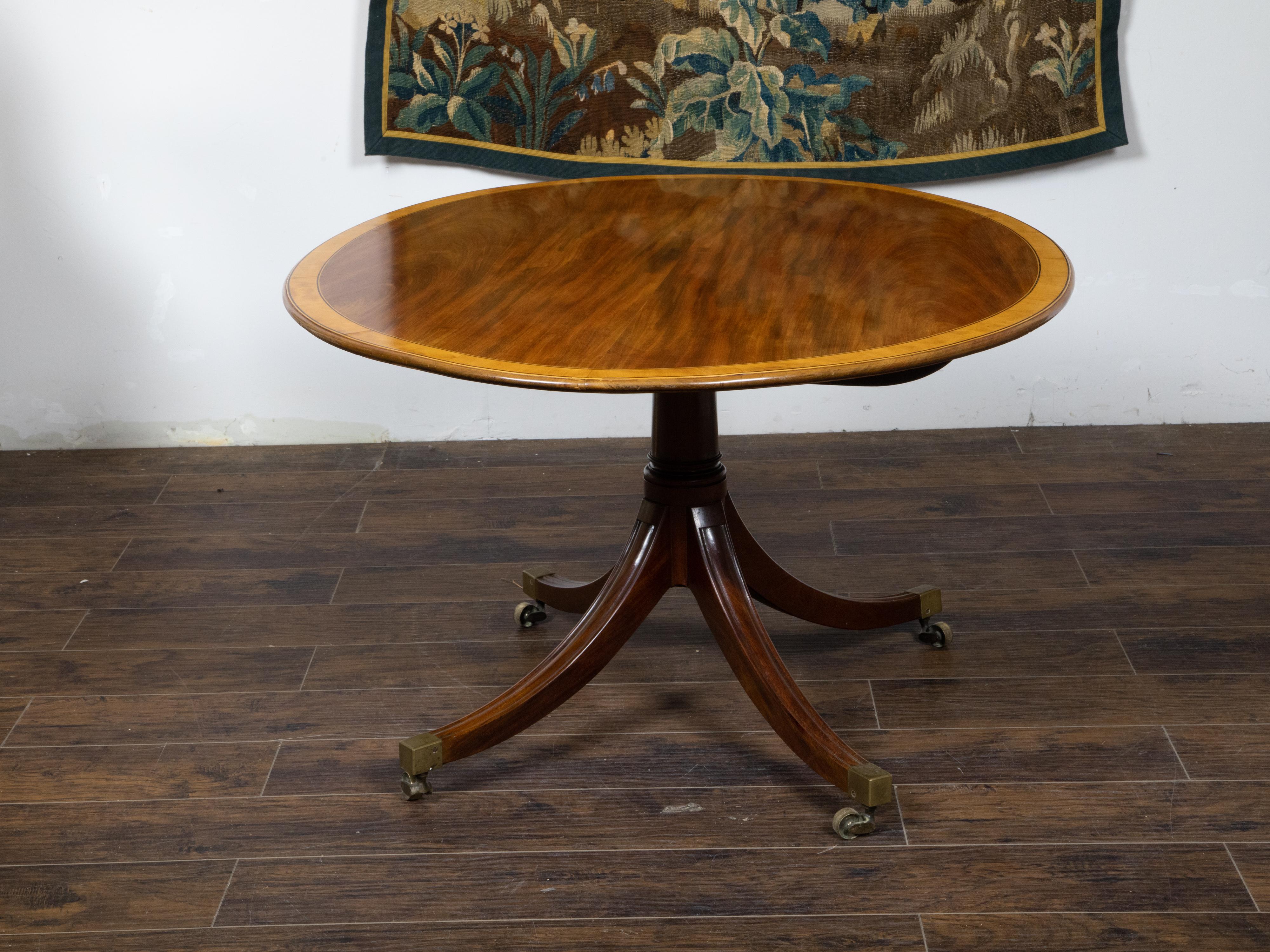 An English mahogany pedestal table from the Mid-19th Century, with oval top, cross banding, quadripod base and brass casters. Created in England during the second quarter of the 19th century, this mahogany table features an exquisite oval top