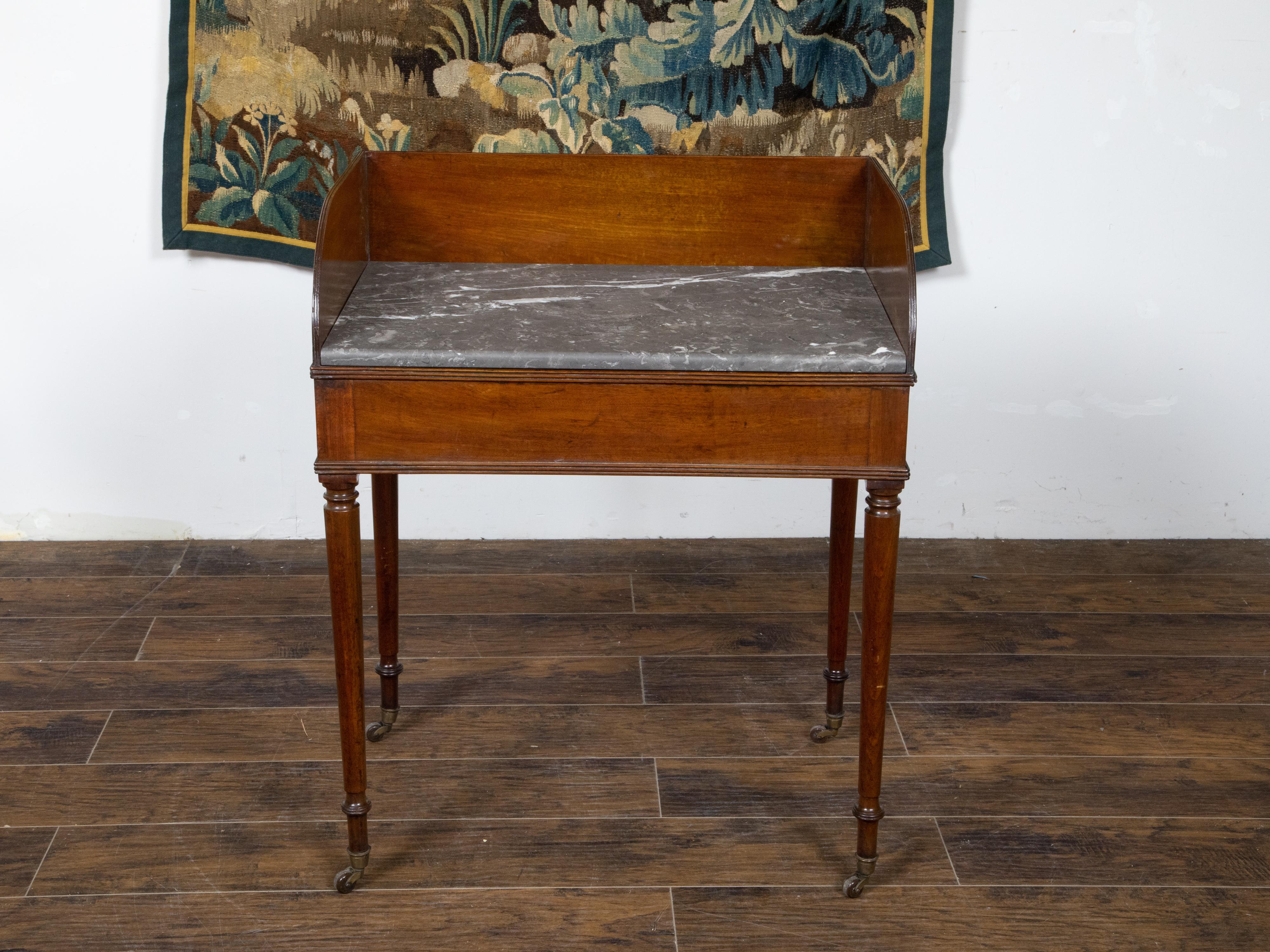 An English mahogany washstand table from the Mid-19th Century, with grey veined marble top, three-quarter frame, turned legs and brass casters. Created in England during the second quarter of the 19th century, this mahogany washstand table features