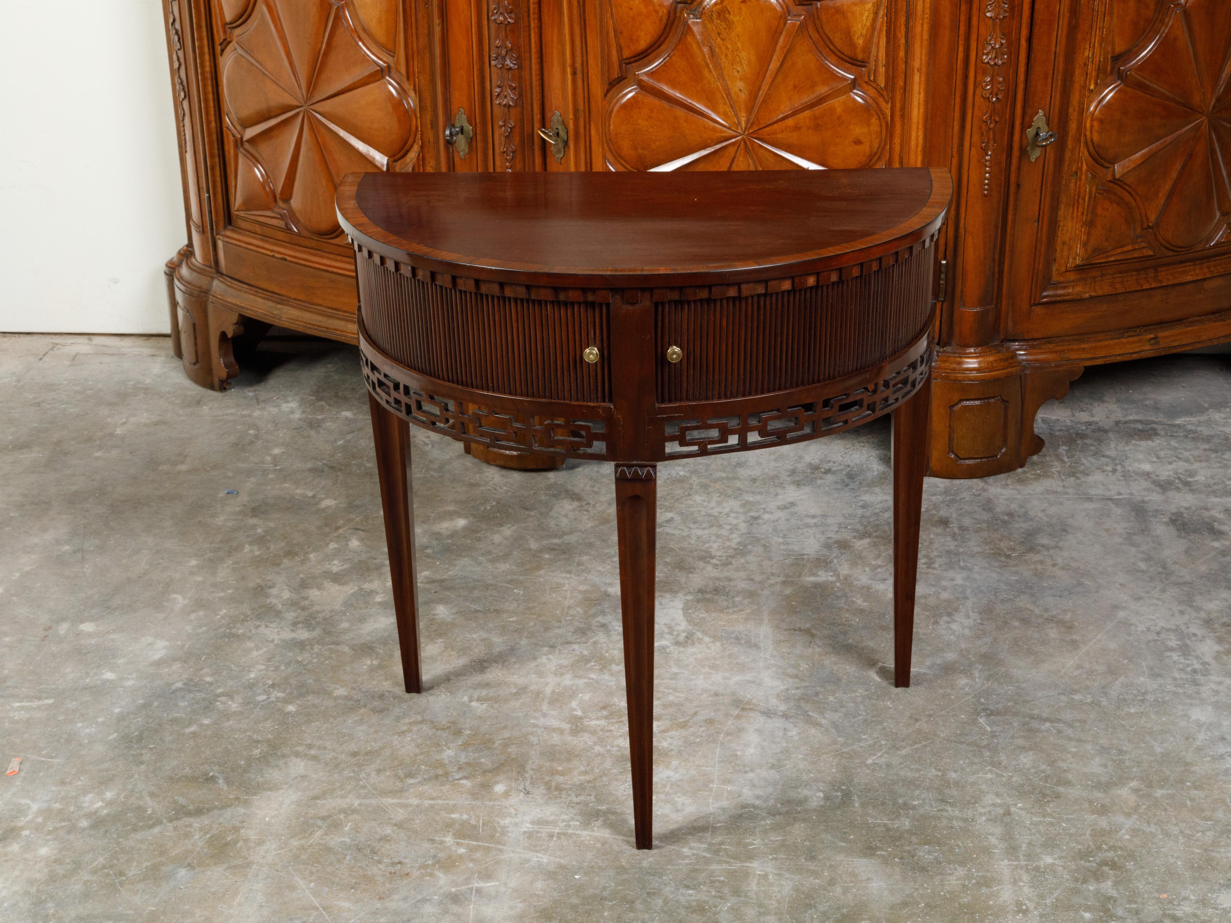 An English mahogany demi-lune console table from the 19th century, with a dentil cornice over two reeded tambour sliding doors and carved apron. Created in England during the 19th century, this demi-lune console table features a semi-circular
