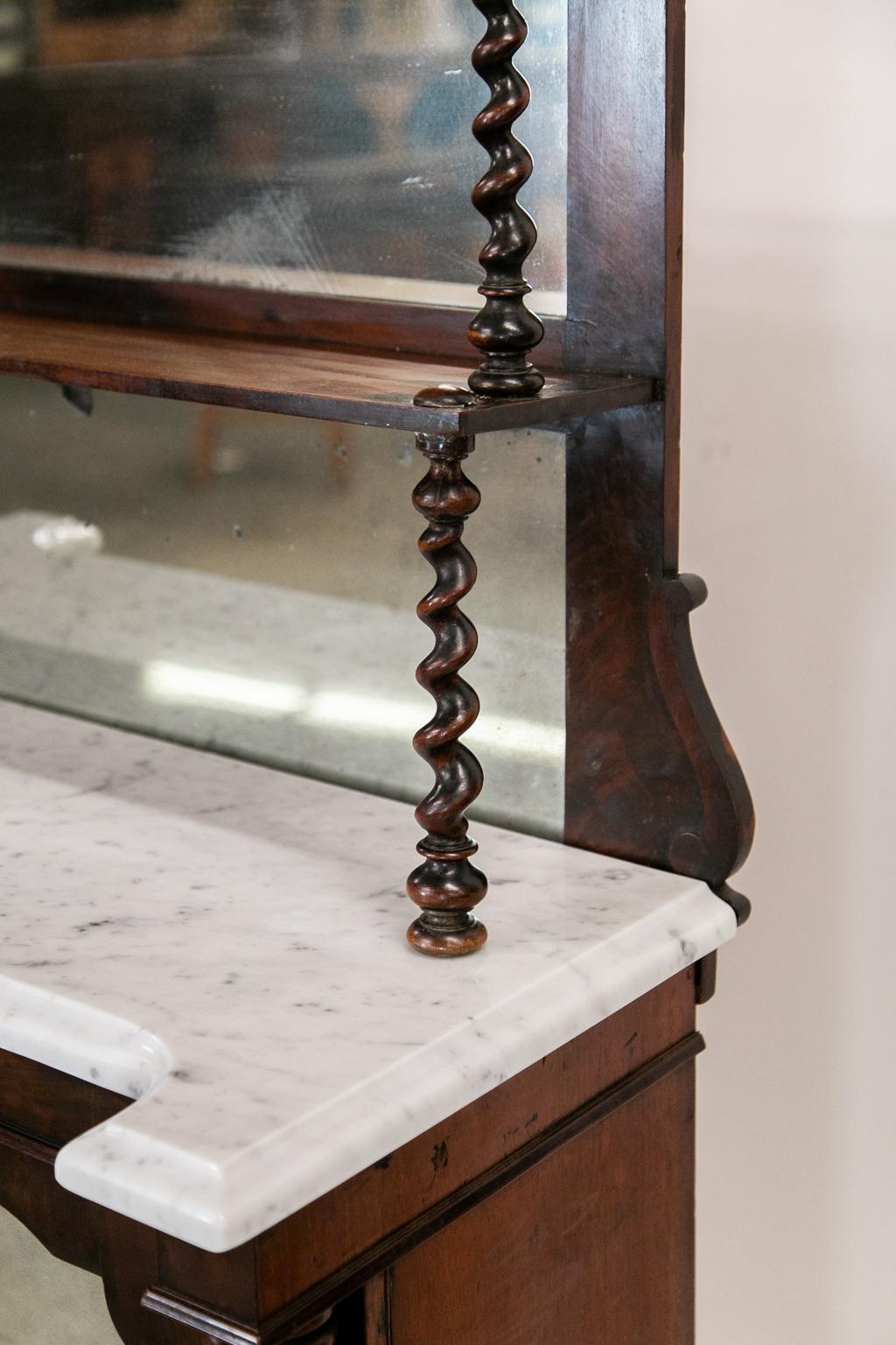 La parte superiore di questo chiffonier in mogano a torsione d'orzo presenta una cresta reticolata ed è sostenuta da quattro colonne a torsione d'orzo. I ripiani e le ante hanno i vetri a specchio originali, tutti rovinati dall'età. La porta