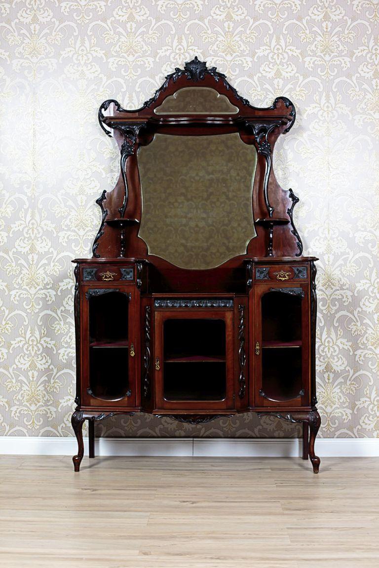 A two-piece cabinet that is composed of a base on high, bent legs, and a setting.
The base is three-door, and is lower in the central part. The door panels are glazed. The inside hides three shelves that are padded with velvet.
To continue, the
