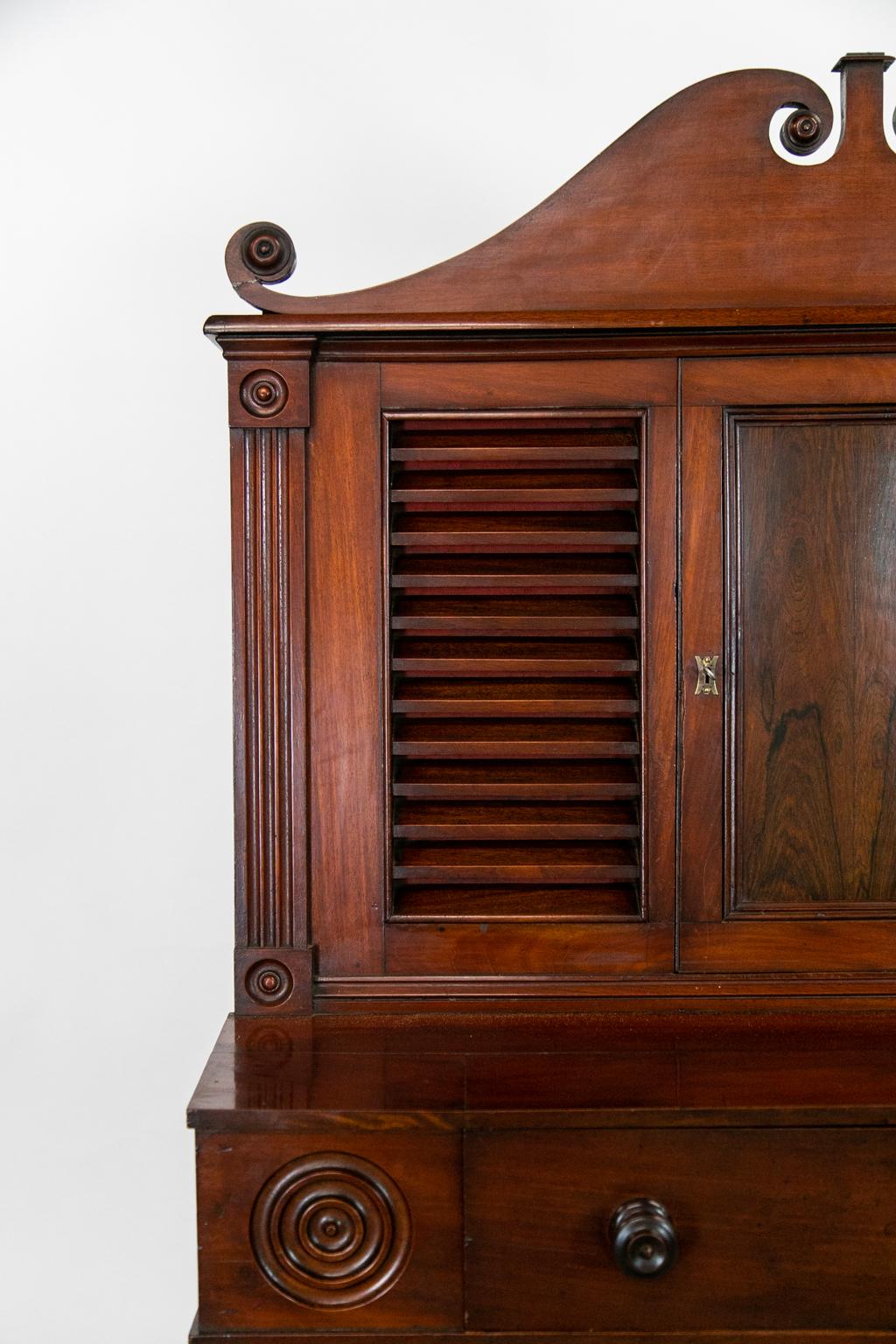 This mahogany cabinet has louvered panels on the front and sides of the top section. The door of the top section has a bookmatched rosewood center panel framed with shaped molding. The top has a broken arch top with turned rosettes at the top and