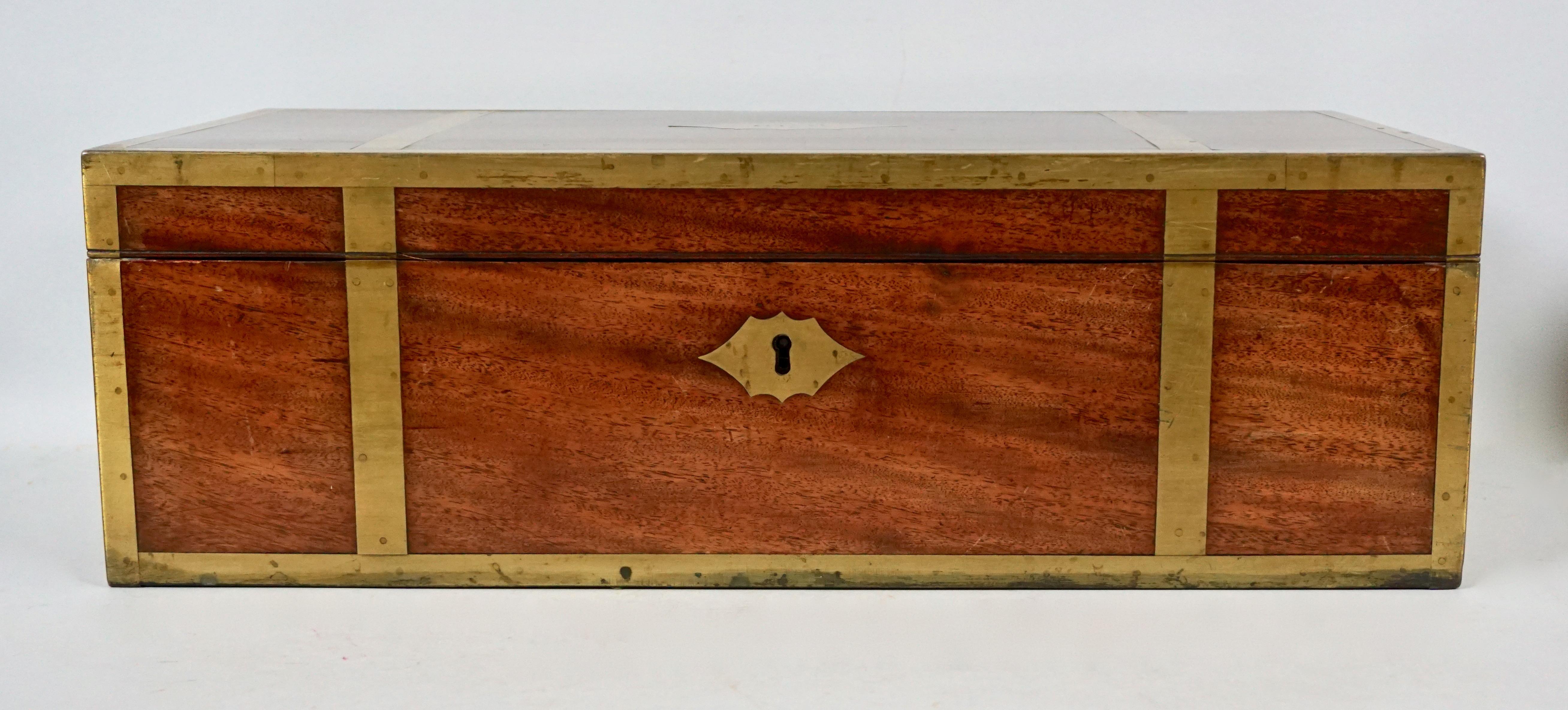 An English mahogany campaign style brass bound traveling desk with recessed handles and a long exterior side drawer, the interior with a velvet lined writing slope, inkwell, pen compartments and letter wells. The top has a cut brass central