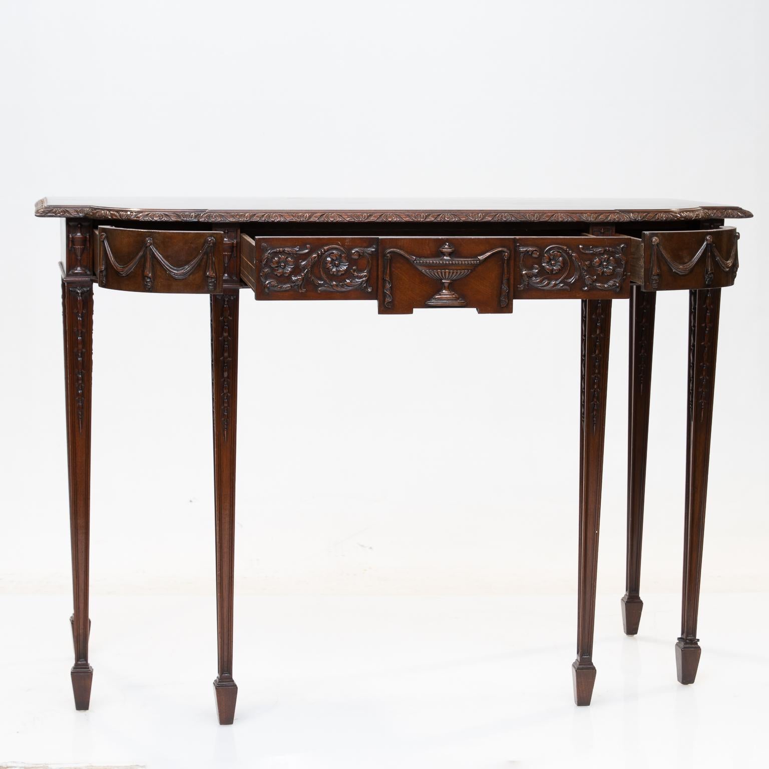 This console is in the style of Robert Adams. English made from choice mahogany wood. Solid mahogany top and carved edge molding. Carved front which reveals three drawers. A larger centre drawer with an urn surrounded with foliage carvings. The two