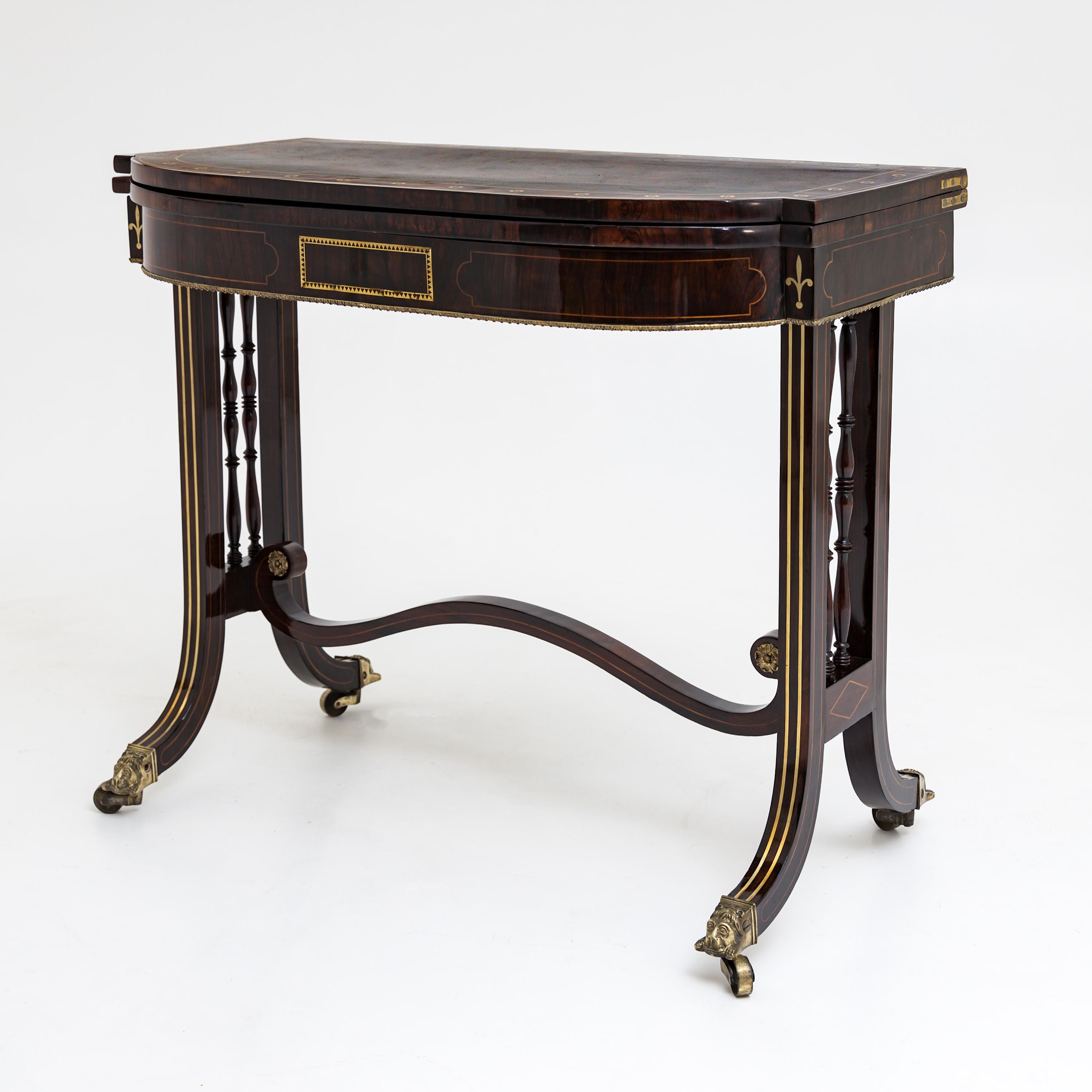 English mahogany console table on wheels with lion heads and brass inlays, turned rods between the legs and a curved intermediate strut. The tabletop is slightly curved at the long sides and covered with green felt on the inside. There is storage