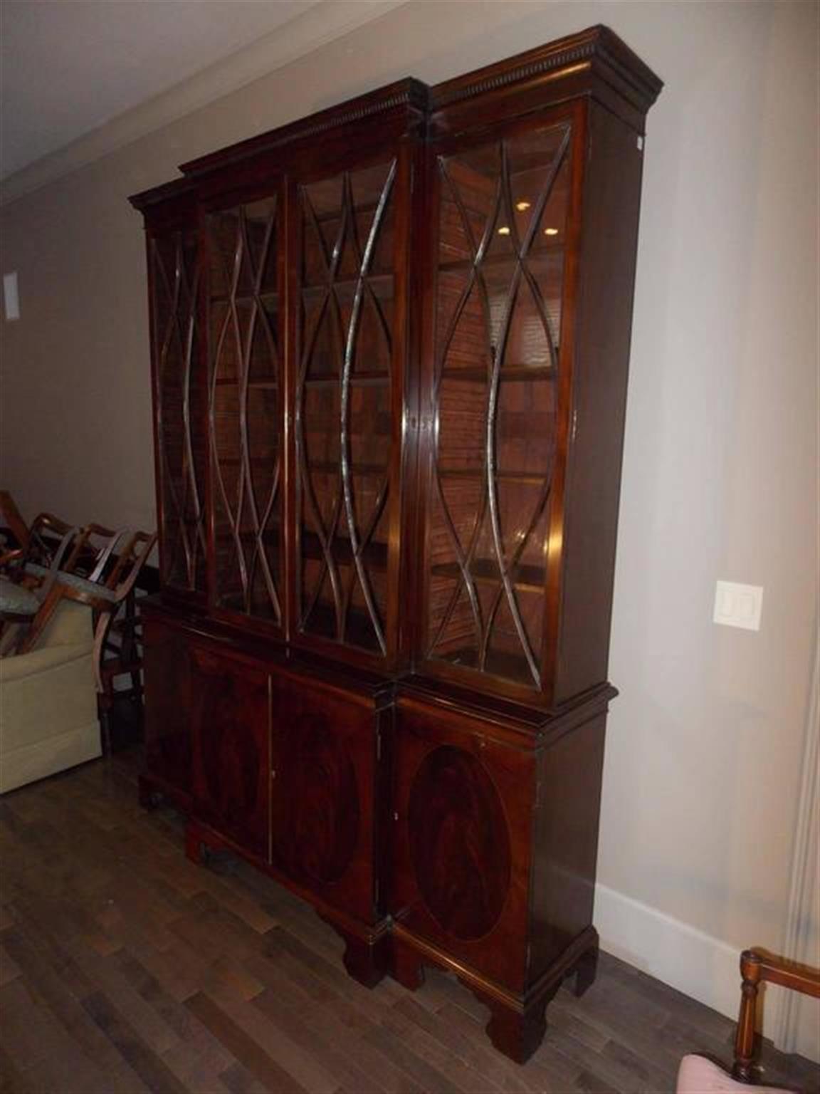English mahogany breakfront with carved dental molding cornice, four upper case glass doors with arched serpentine mullions, adjustable interior shelving, flanking lower case oval inlaid cabinets concealing interior shelving and resting on a raised