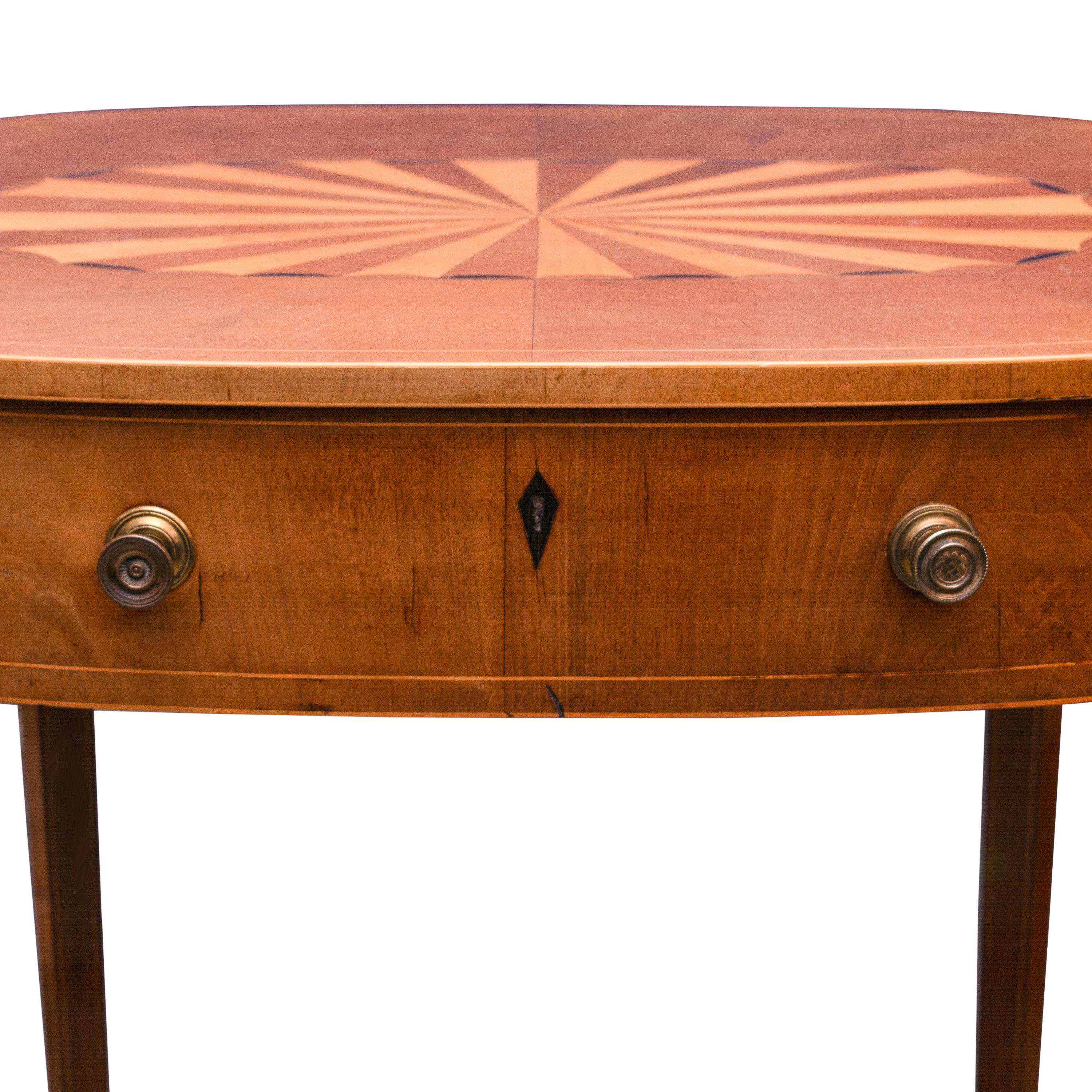 Il s'agit d'un meuble ovale traditionnel anglais en acajou clair, dont le dessus est incrusté de marqueterie en forme de tarte, au-dessus d'une large frise contenant un tiroir central avec un écusson en ébène. La partie supérieure est soutenue par