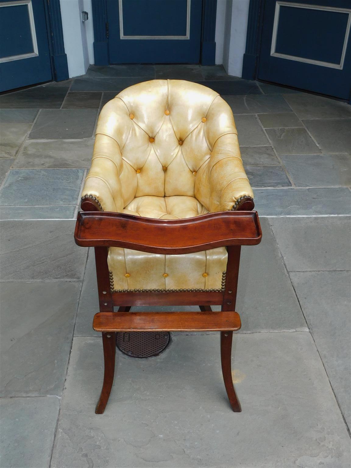 English Mahogany leather tufted and brass tacked arched high chair with upper removable tray, lower adjustable and removable leg rest, and resting on the original flanking four splayed legs, Early 19th Century.