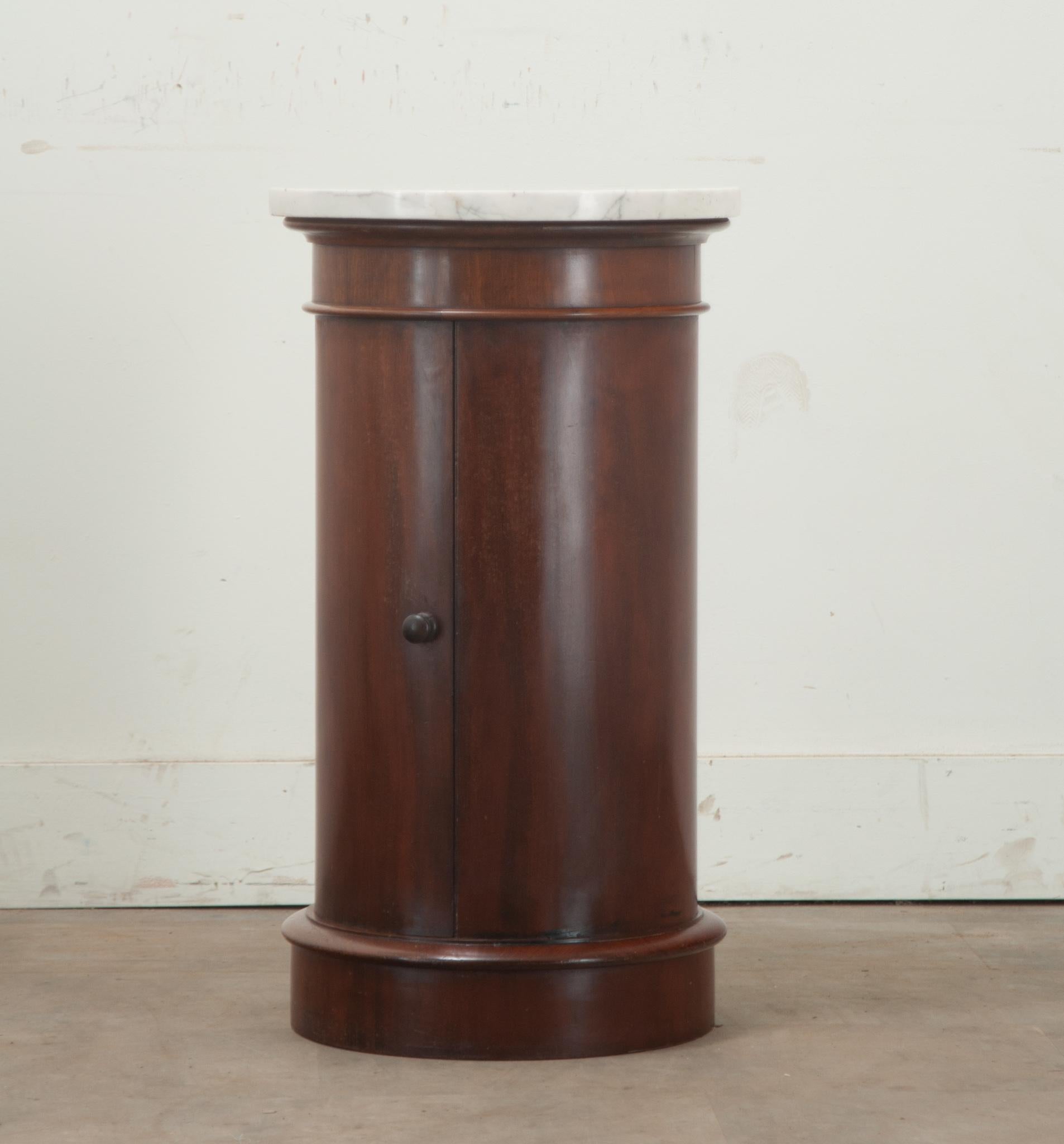 A round mahogany and marble English bedside table. This table has a white beveled marble top over a mahogany body with a door that has a turned knob and opens to reveal a single fixed circular shelf at 10 ½'' deep. The whole sits on a round plinth