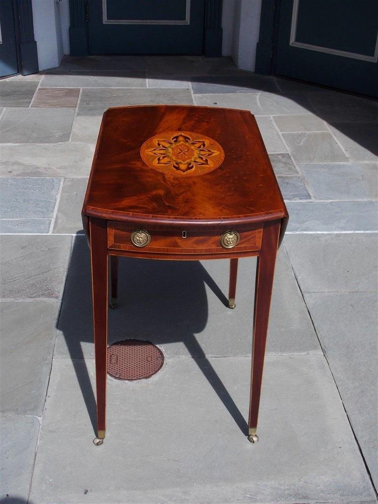 English mahogany oval foliage satinwood inlaid one drawer drop-leaf pembroke table cross banded in tulip wood, flanking original brasses, and terminating on tapered legs with the original cup barrel casters. Late 18th century. 

Pembroke table is