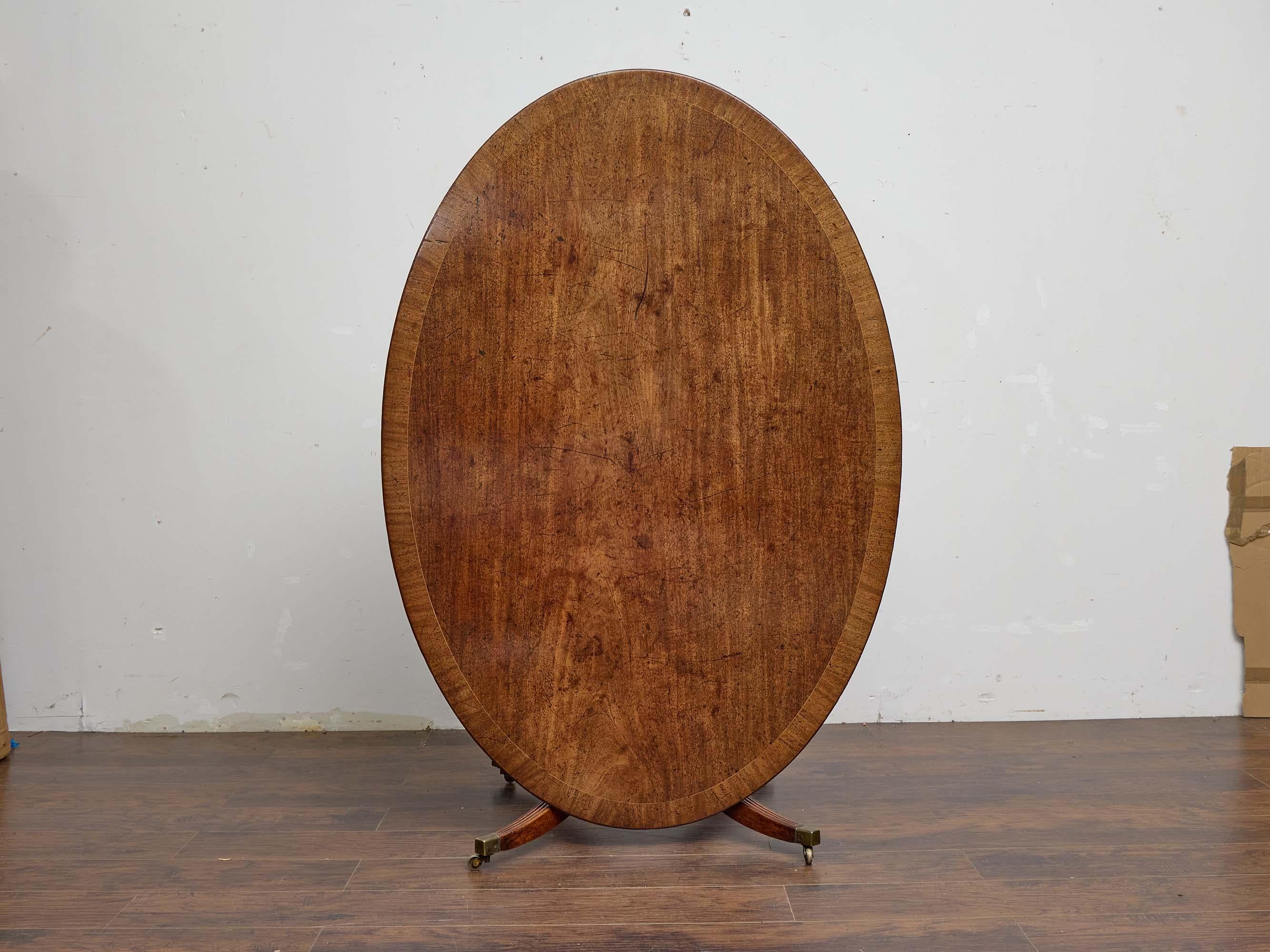 Esta mesa inglesa con tablero basculante de principios del siglo XIX ejemplifica la refinada artesanía de la época de la Regencia en caoba maciza. La parte superior ovalada presenta una cálida pátina y está enmarcada con un fino rebordeado, que