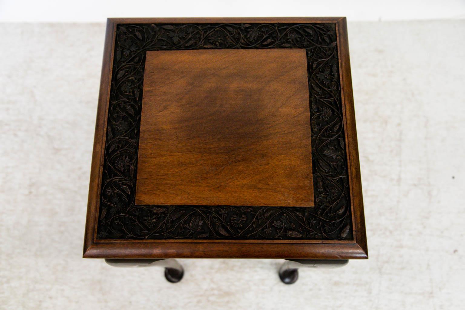 The center of this table has a square mahogany panel framed with an intricately carved border of interlaced leaves, vines, and flowers. The four sided table has cabriole legs that terminate in pad feet.