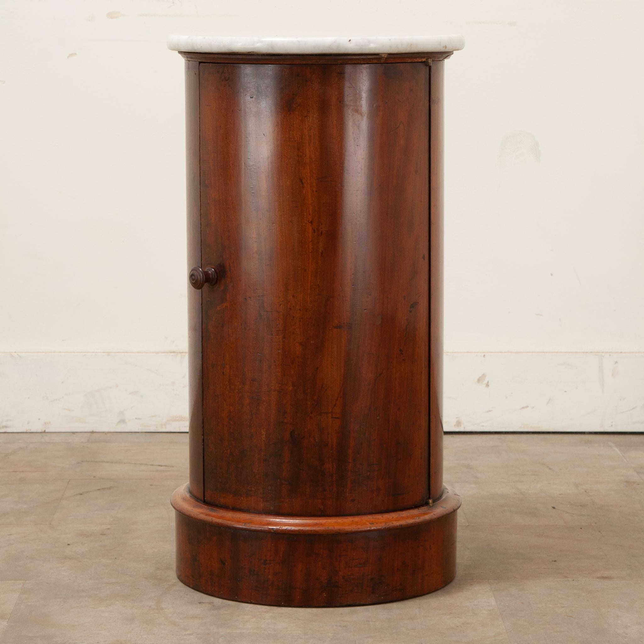 A round mahogany and marble English bedside table. This table has a white beveled marble top over a mahogany body with a door that has a turned knob and opens to reveal a single circular fixed shelf at 10 ¾” deep. The whole sits on a round plinth