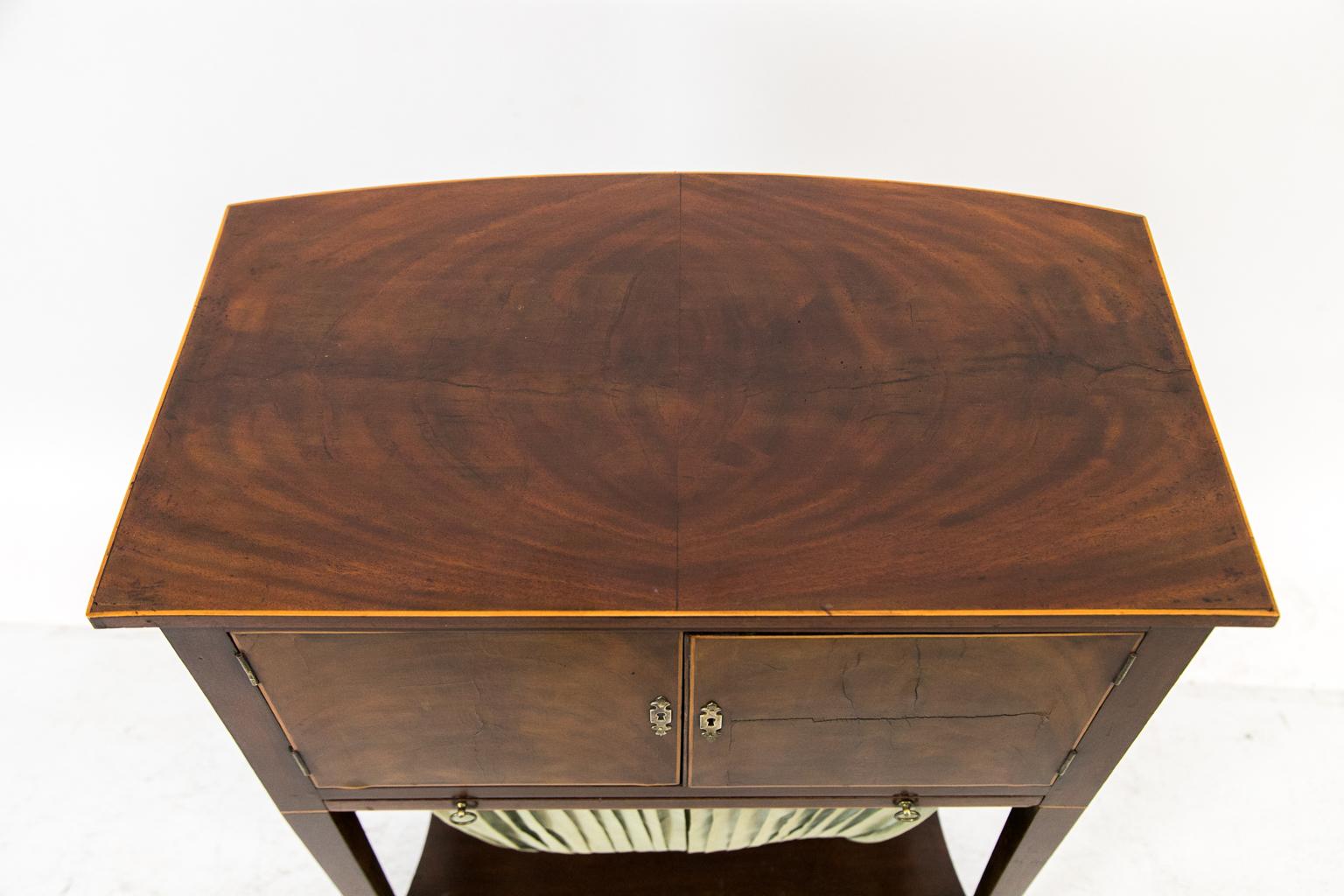 This sewing table has a bookmatched flame mahogany top with inlaid boxwood edging. The doors have flame grained mahogany also with boxwood edging. The right-hand door has a shrinkage crack in the middle. The pull-out yarn basket has pleated green