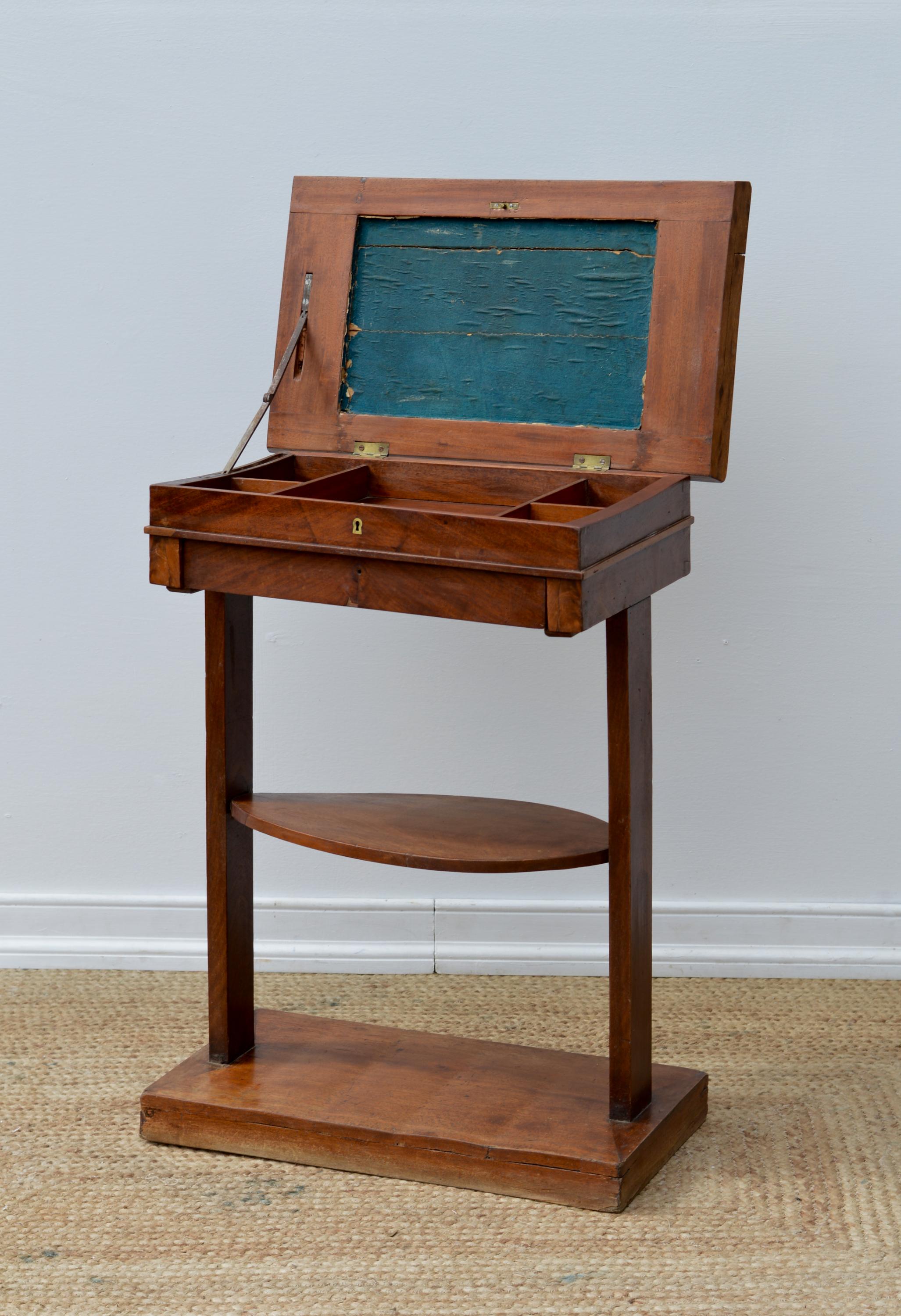 A fetching and petite writing stand of mahogany having a lift and hinged top revealing slotted compartments above a single slide out drawer perched atop a floating oval gallery that terminates upon simple and rectangular stiles and a conforming