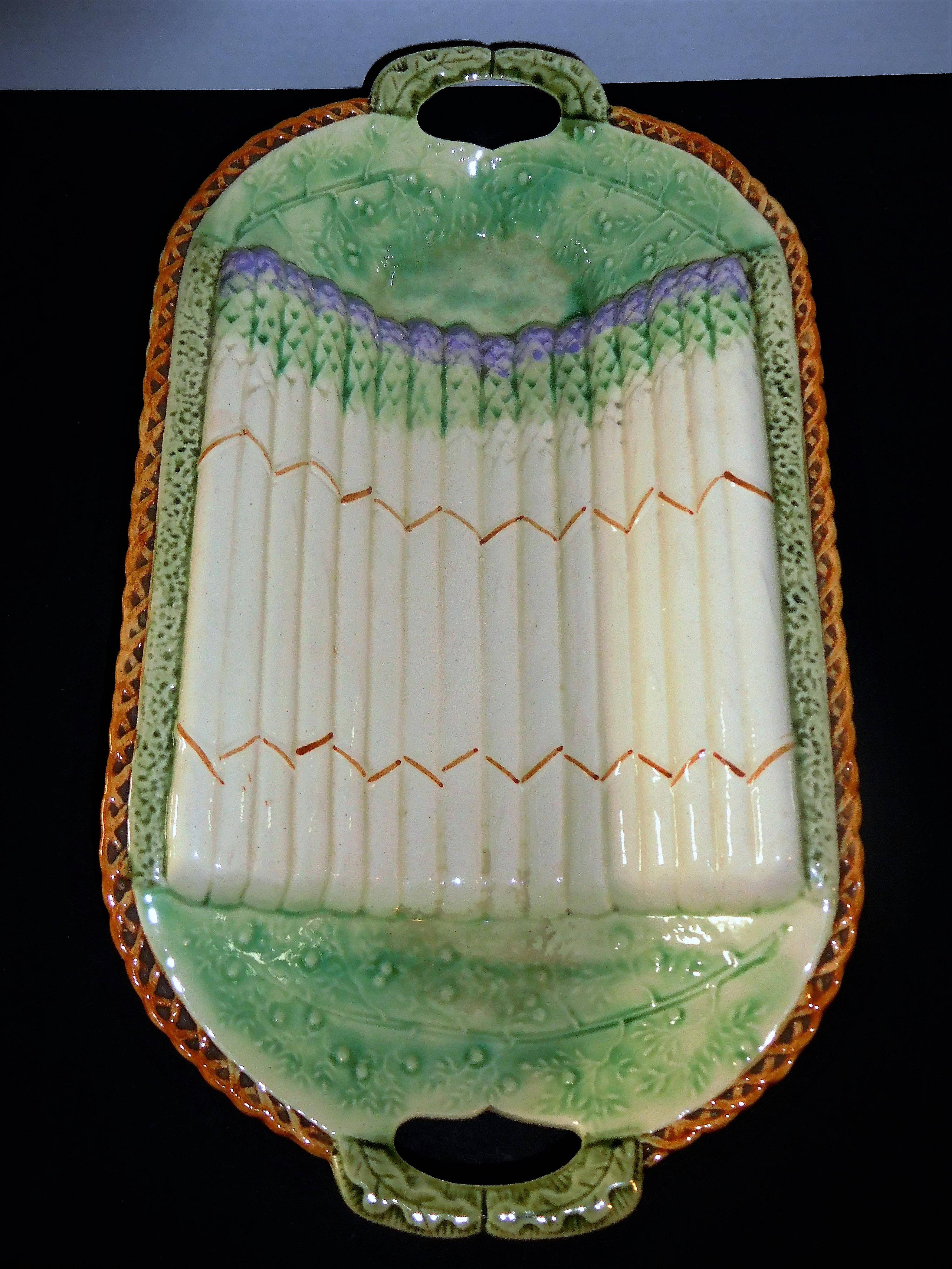 This majolica asparagus cradle resting atop an attached tray with pierced handles, has a brown basket-weave edge and four brown branches as feet. The interior of the basket is light green with bas-relief decoration of stylized asparagus stems, and