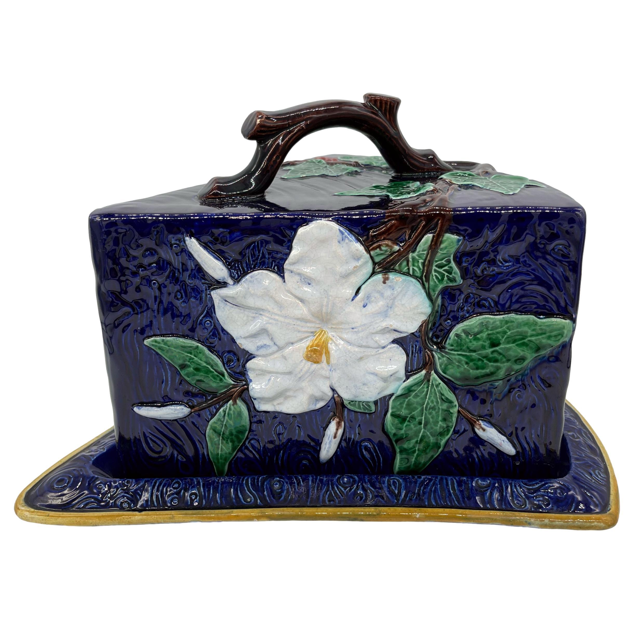 Large Majolica cheese keeper and stand, molded in the shape of a cheese wedge wrapped in vines, green glazed ivy leaves with red berries, and relief-molded trompe l'oeil large white flowers, on a faux bois cobalt blue-glazed ground, surmounted with