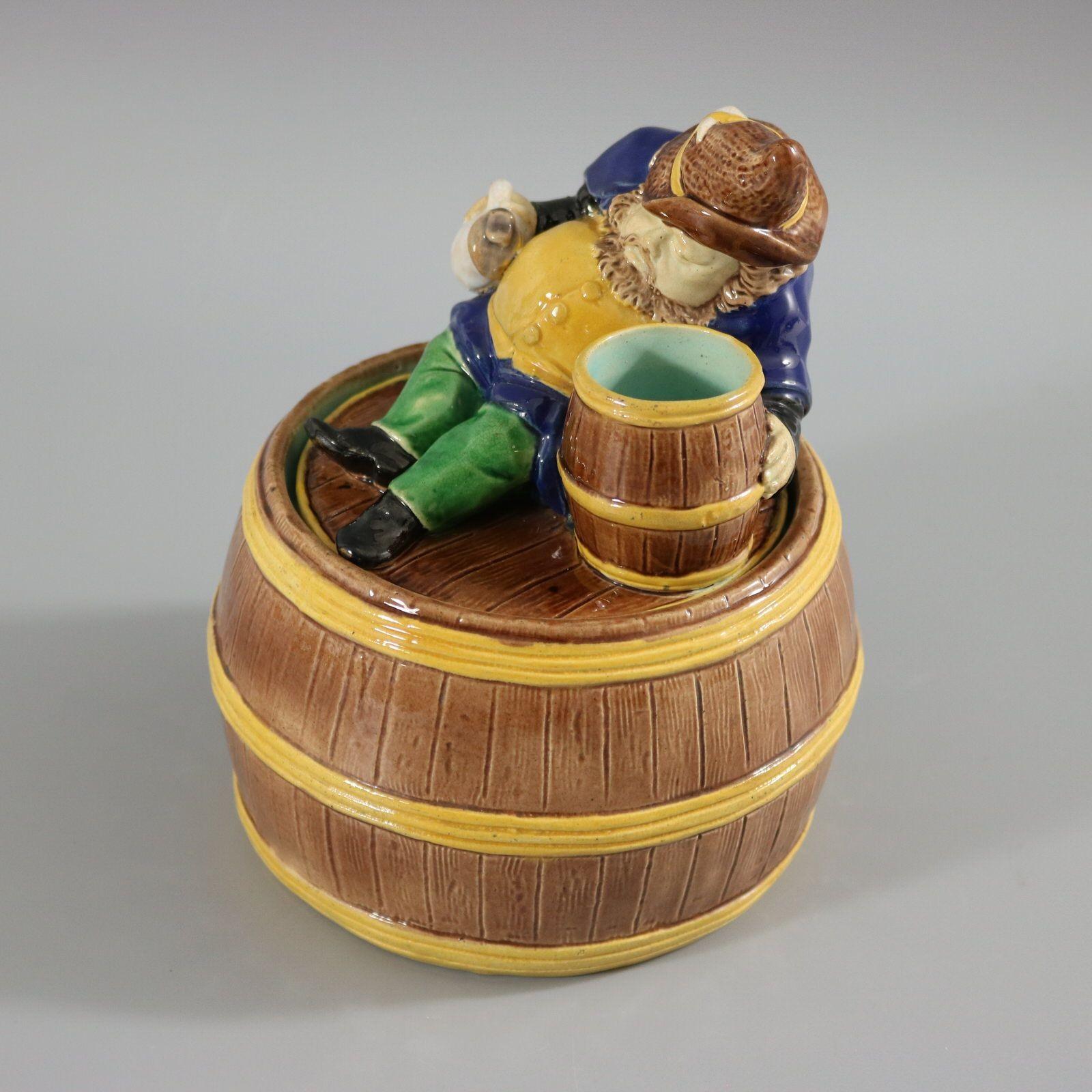 Pot et couvercle en majolique anglaise non identifiés représentant un gnome (ou un nain) allongé à côté d'un tonneau de bière, avec une cruche dans sa main droite. La base a la forme d'un tonneau. Coloration : le brun, le jaune, le bleu cobalt, sont