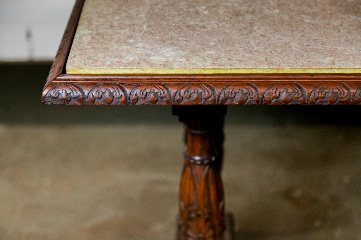 The top edge of this center table has a carved and molded shape. The base has four support columns that have carved fluting and floral medallions. The side apron has a carved plume with a stylized pineapple pendant below. The columns end in a