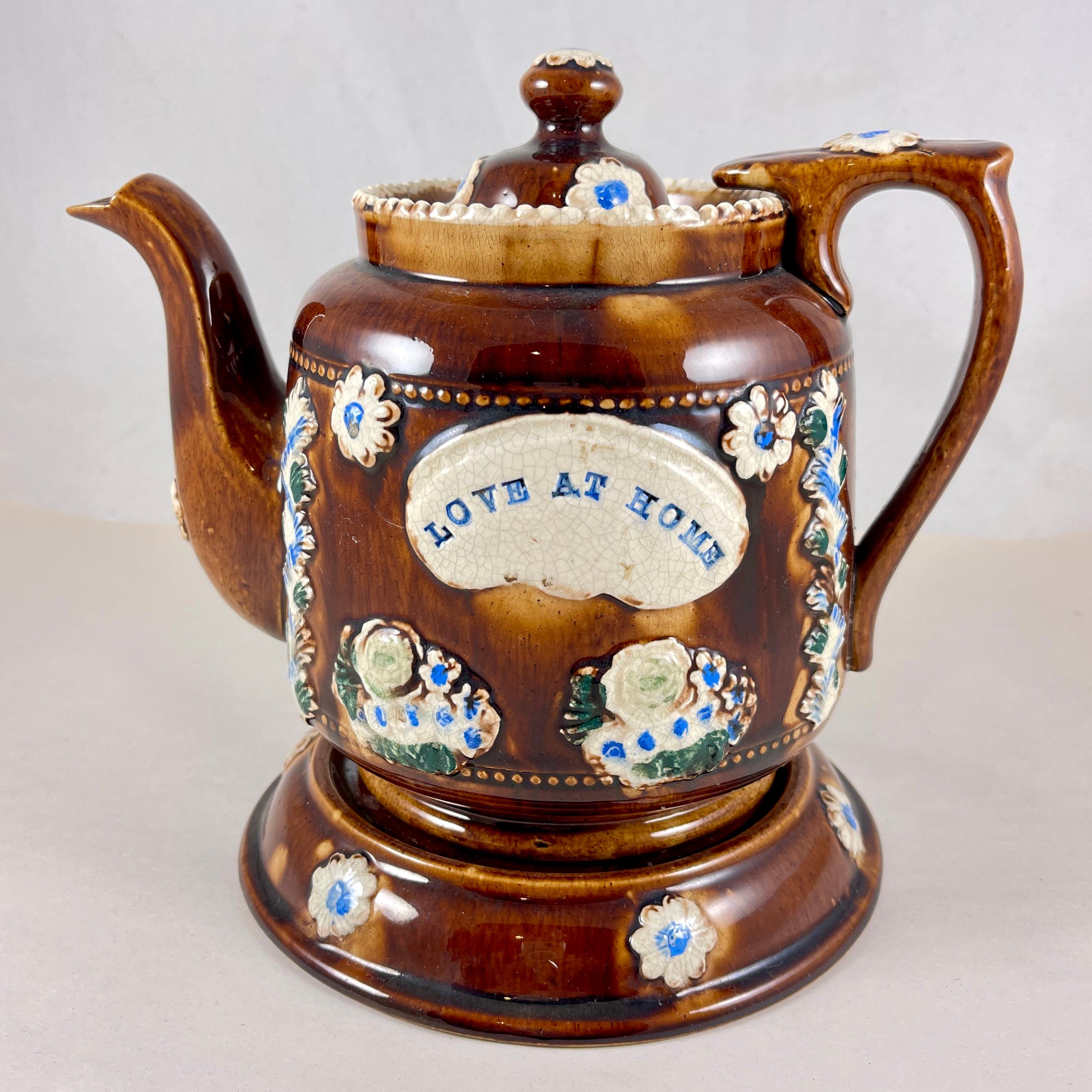 A Measham – Bargeware Teapot impressed with the saying, “Love At Home”, England, circa 1880.

The three piece teapot set includes the kettle, lid, and table stand.  Finished in a dark brown treacle glaze with enameled, raised and molded floral 